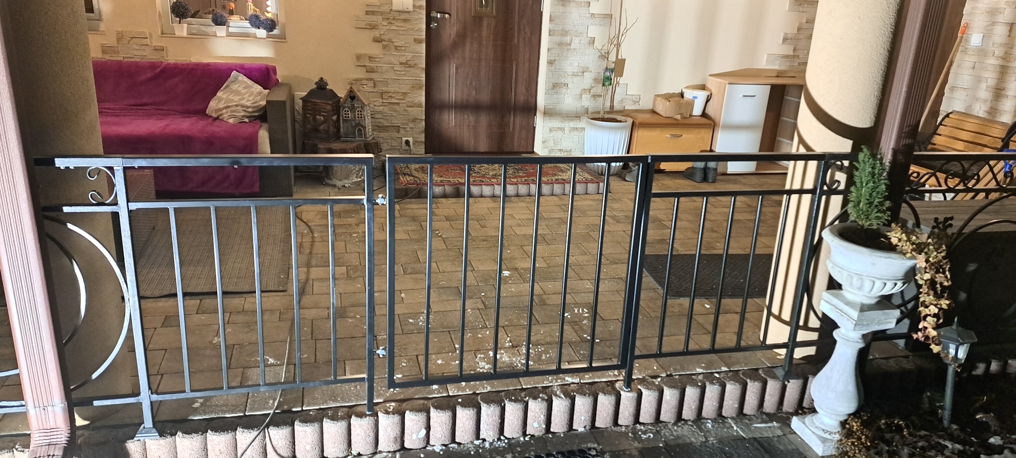 Czarna, metalowa konstrukcja z pionowymi prętami, tworząca zabezpieczenie tarasu. W tle widoczny fragment wnętrza domu z meblami i dekoracjami.