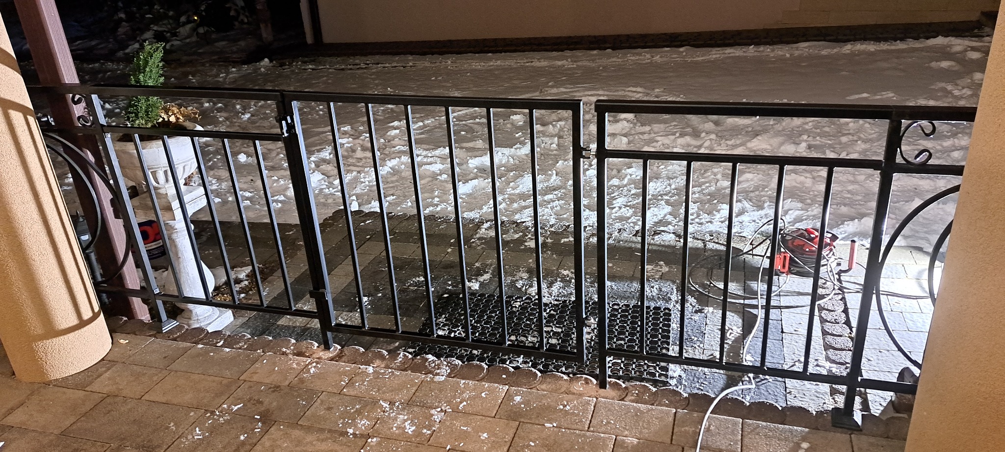 Czarne, metalowe ogrodzenie z furtką, widok z bliska na tle ośnieżonego terenu. Dekoracyjny element w górnej części słupka. Widoczny kabel elektryczny.