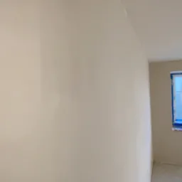 Ściana po świeżo położonej gładzi, widoczne zabezpieczone okno folią malarską. Gładka powierzchnia gotowa do malowania, dająca wrażenie czystości i minimalizmu.