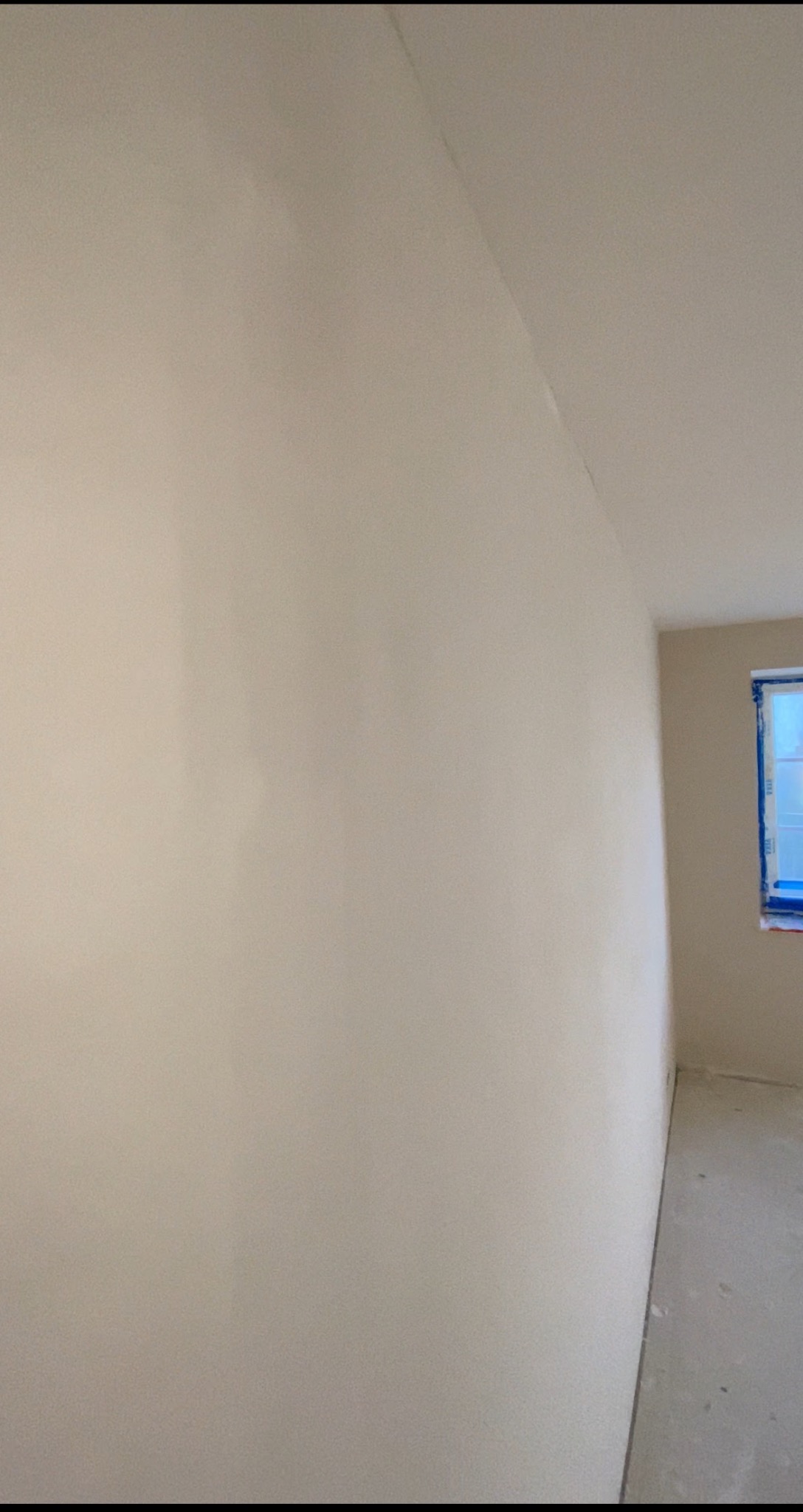 Ściana po świeżo położonej gładzi, widoczne zabezpieczone okno folią malarską. Gładka powierzchnia gotowa do malowania, dająca wrażenie czystości i minimalizmu.