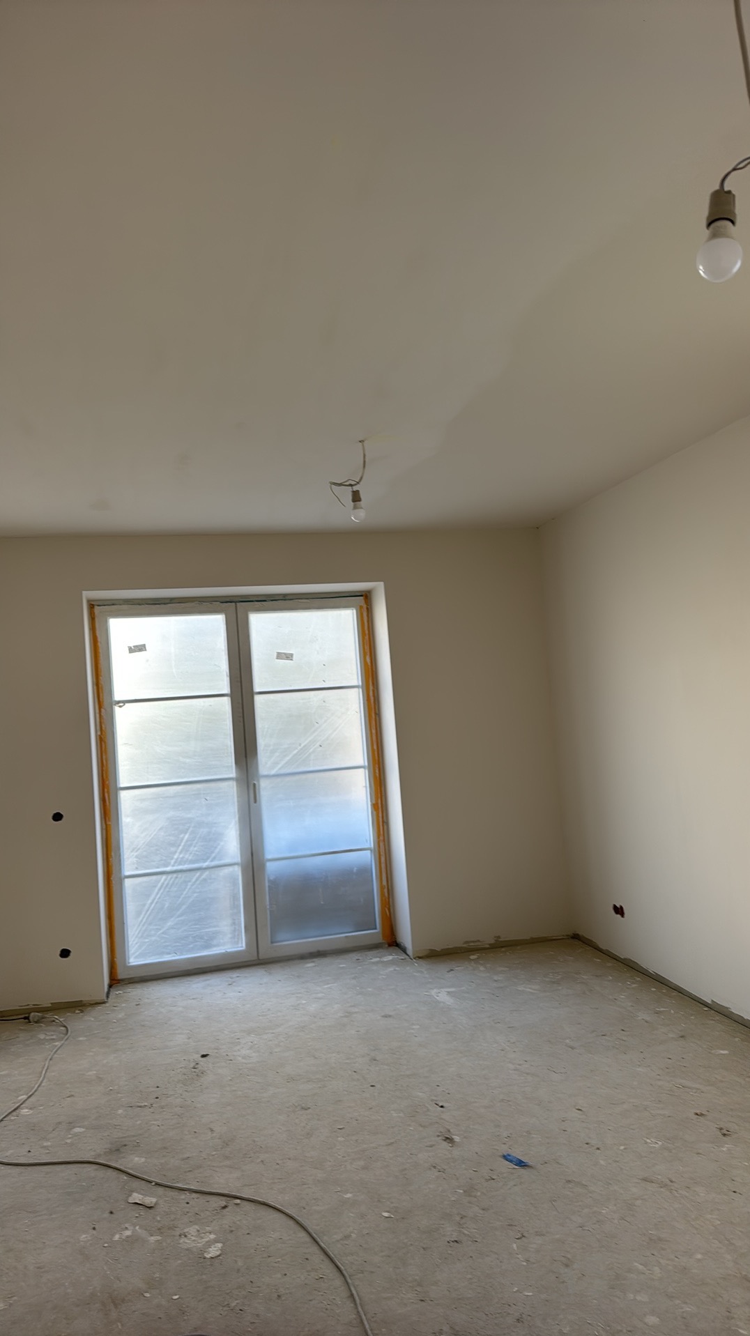 Pokój w trakcie remontu: świeżo wyszpachlowane ściany, okno zabezpieczone folią, surowa podłoga. Widoczne niedoskonałości i przewody elektryczne.