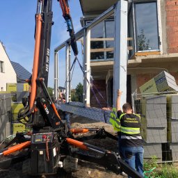 PRZEDSIĘBIORSTWO HANDLOWO-USŁUGOWE MAGDALENA MATUSIAK - Montaż okna z użyciem dźwigu na budowie. Pracownicy w kamizelkach odblaskowych. Widoczne bloczki Lubau. Częściowo zamontowane okno na wyższej kondygnacji.