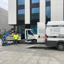 PRZEDSIĘBIORSTWO HANDLOWO-USŁUGOWE MAGDALENA MATUSIAK - Montaż okien w nowoczesnym budynku. Pracownicy z wózkiem transportowym i samochodem dostawczym firmy Matusiak. Widoczne okna i reklama rolet na samochodzie.