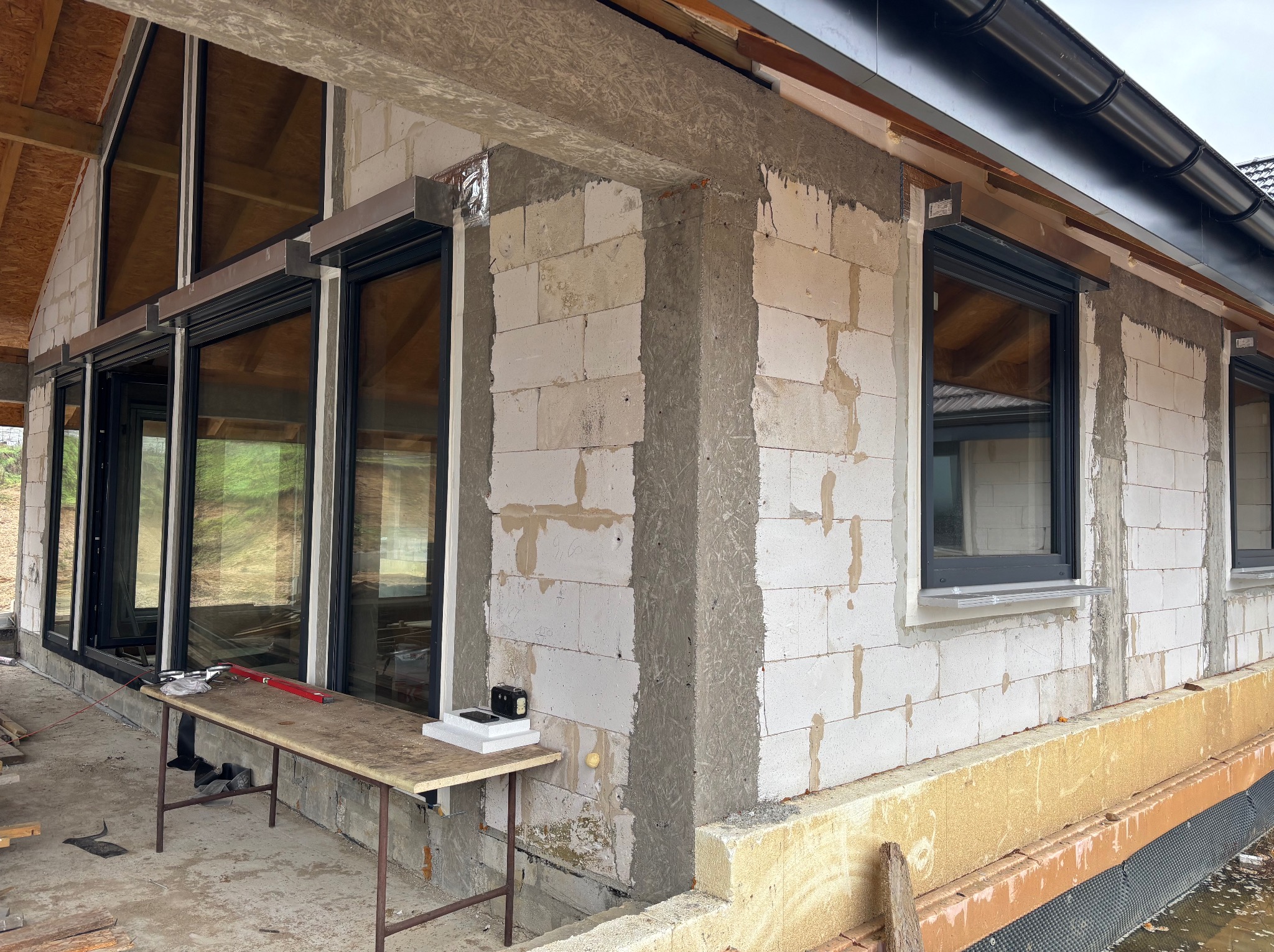 Dom w trakcie budowy z zamontowanymi oknami z roletami zewnętrznymi. Widoczne materiały budowlane i narzędzia na zewnątrz budynku.