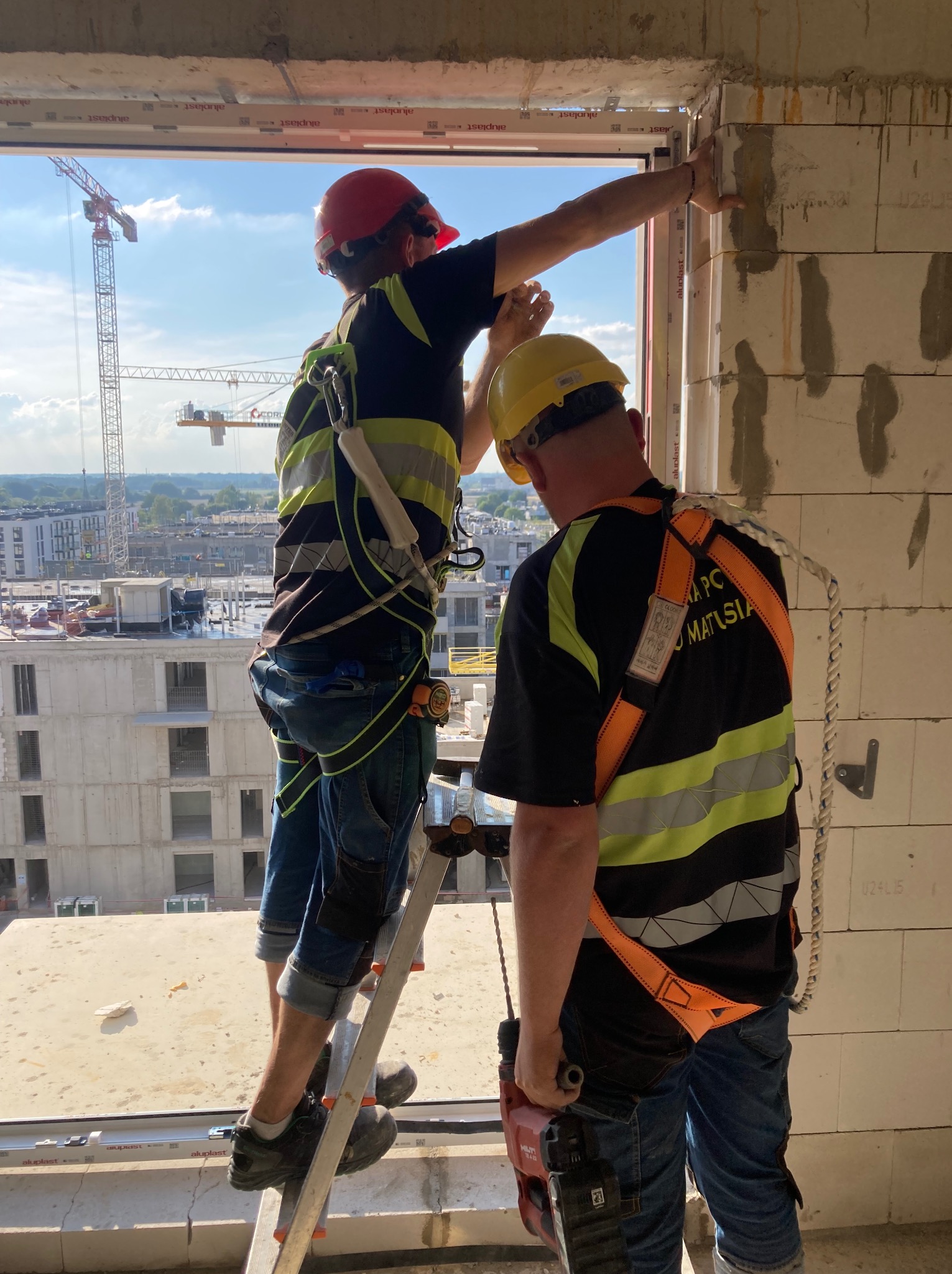 Dwóch monterów w kaskach instaluje okno na budowie. Jeden stoi na drabinie, drugi trzyma wiertarkę. W tle widok na miasto i żurawie budowlane.