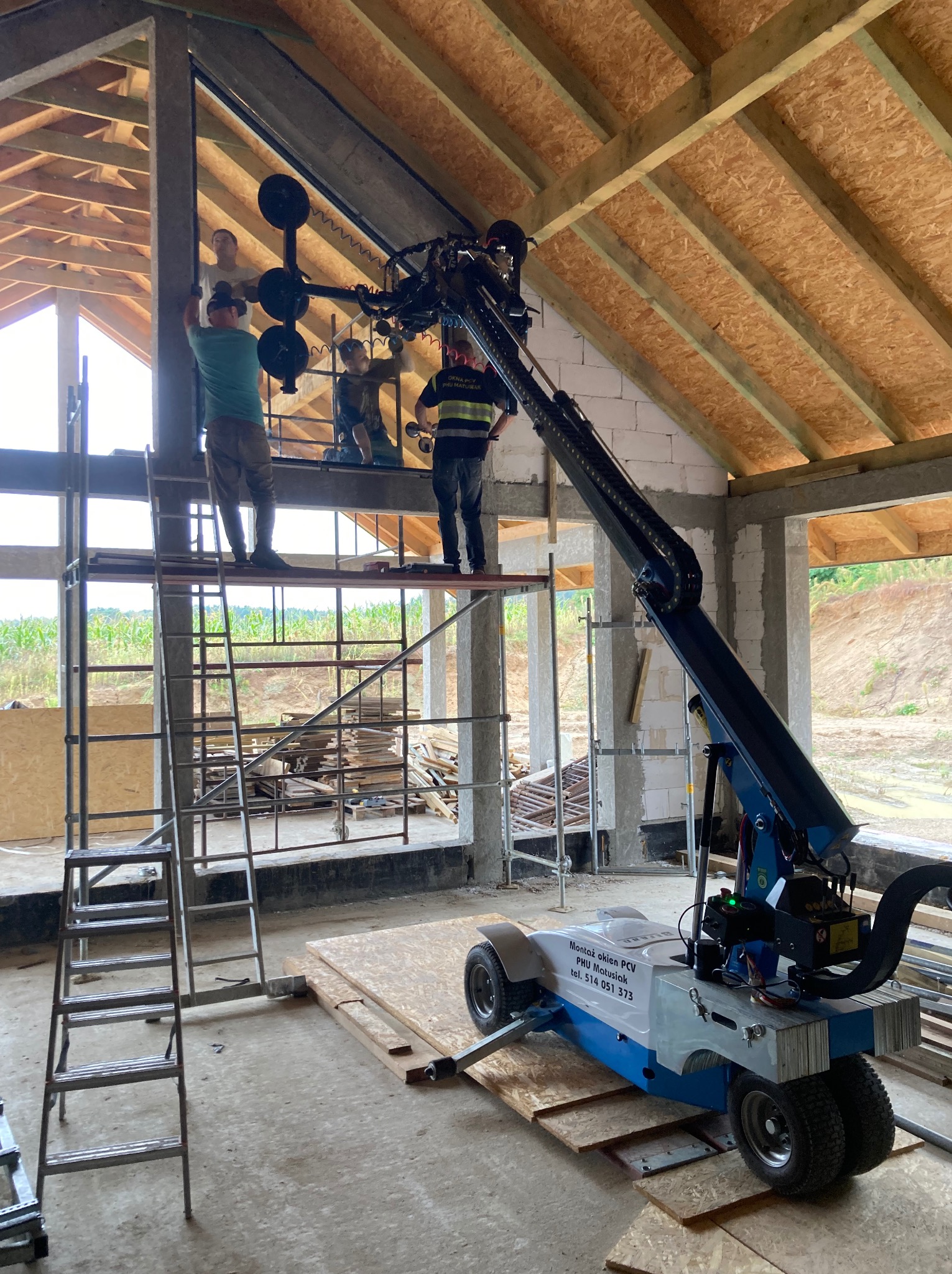 Montaż okna PCV w surowym budynku z użyciem specjalistycznego podnośnika. Pracownicy na rusztowaniu. Widoczne elementy konstrukcyjne dachu i ścian.