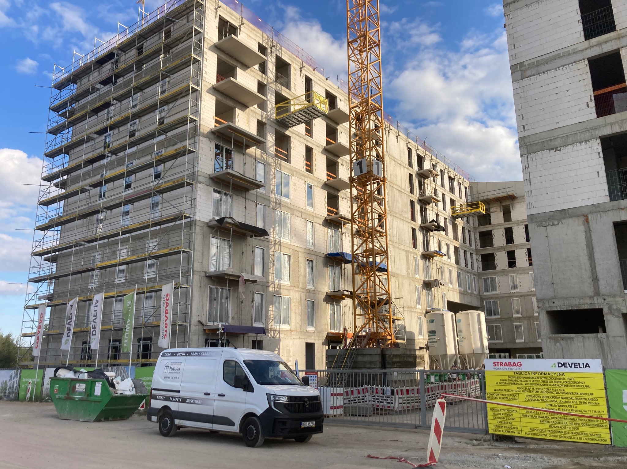 Budowa bloku mieszkalnego w trakcie realizacji, widoczne rusztowania, dźwig i samochód dostawczy z reklamą okien, drzwi i rolet. Tablica informacyjna budowy na pierwszym planie.