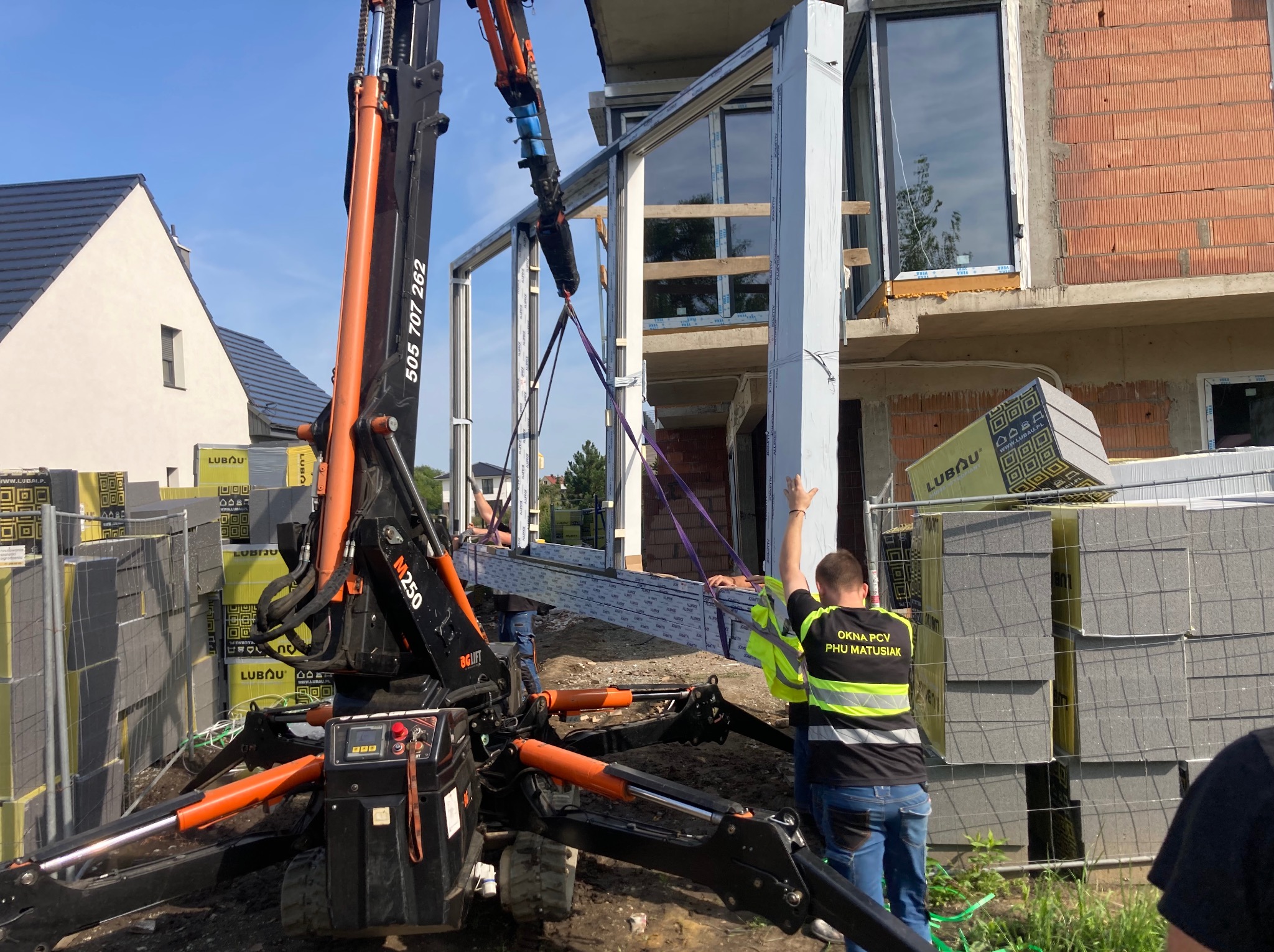 Montaż okna z użyciem dźwigu na budowie. Pracownicy w kamizelkach odblaskowych. Widoczne bloczki Lubau. Częściowo zamontowane okno na wyższej kondygnacji.