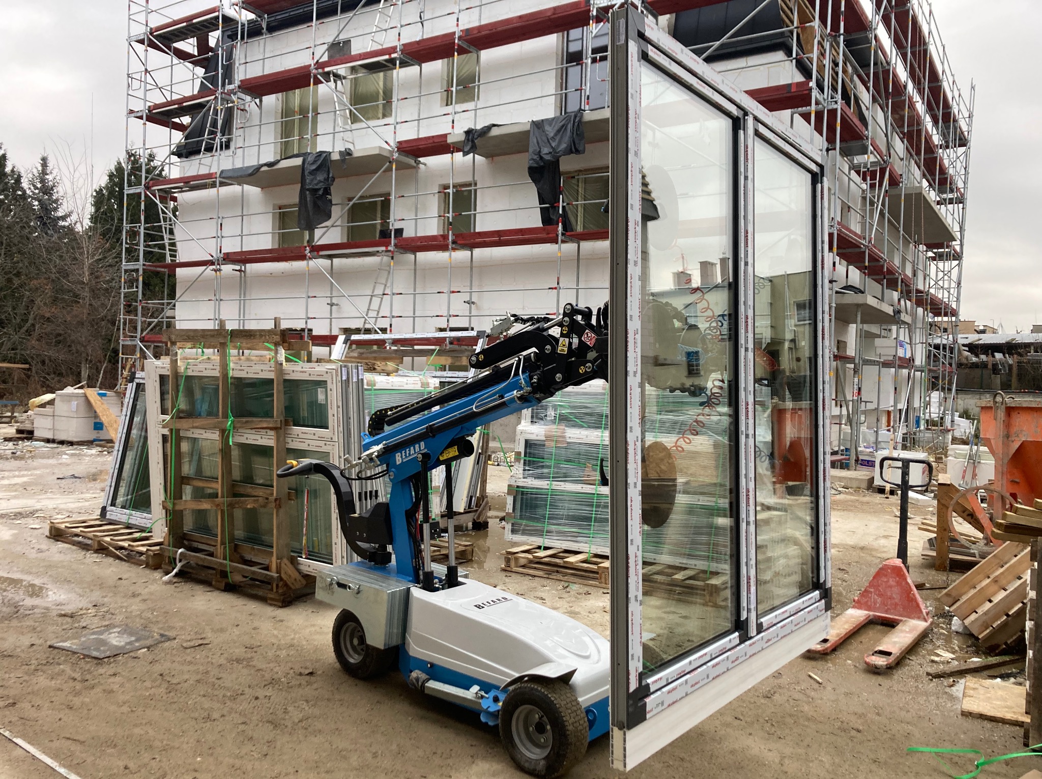 Montaż okna na budowie z użyciem specjalistycznego podnośnika. W tle rusztowanie i okna czekające na montaż. Widoczne elementy zabezpieczające.