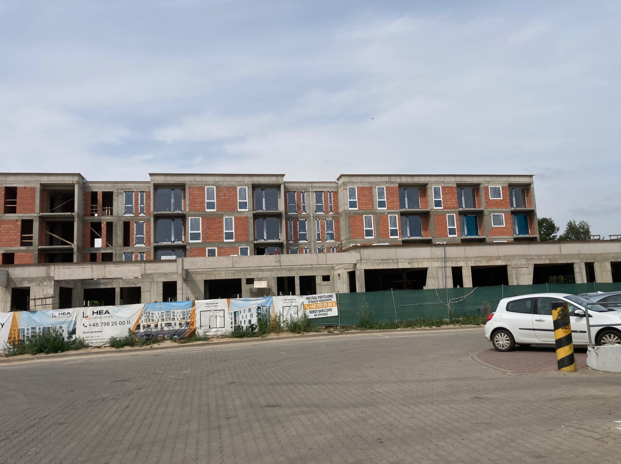 Budynek w stanie surowym z zamontowanymi oknami, widoczne banery reklamowe dewelopera, samochód na parkingu. Ujęcie z poziomu ulicy.