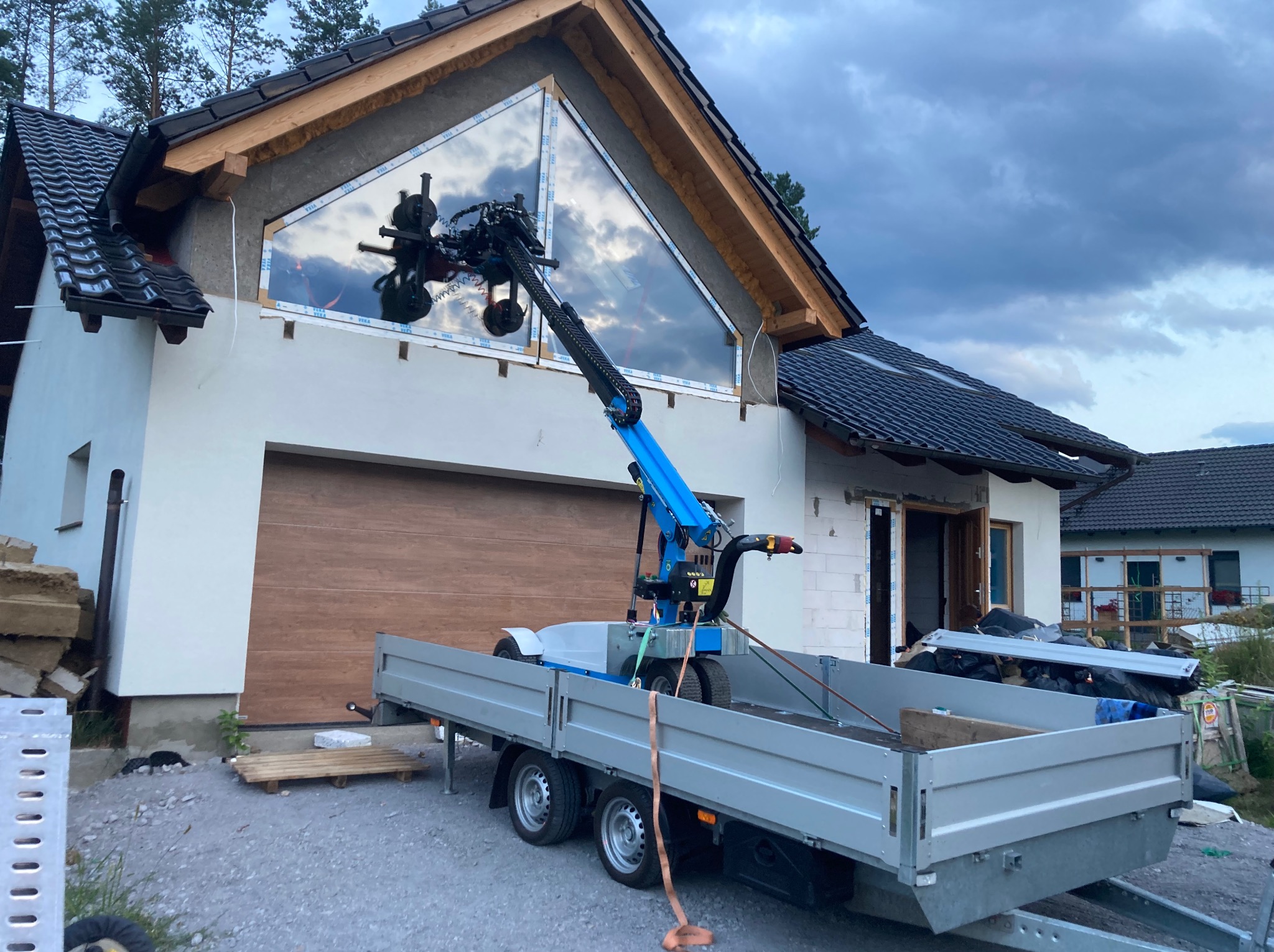 Montaż okna o nietypowym kształcie za pomocą specjalistycznego dźwigu na przyczepie. Widoczny surowy stan elewacji budynku jednorodzinnego.