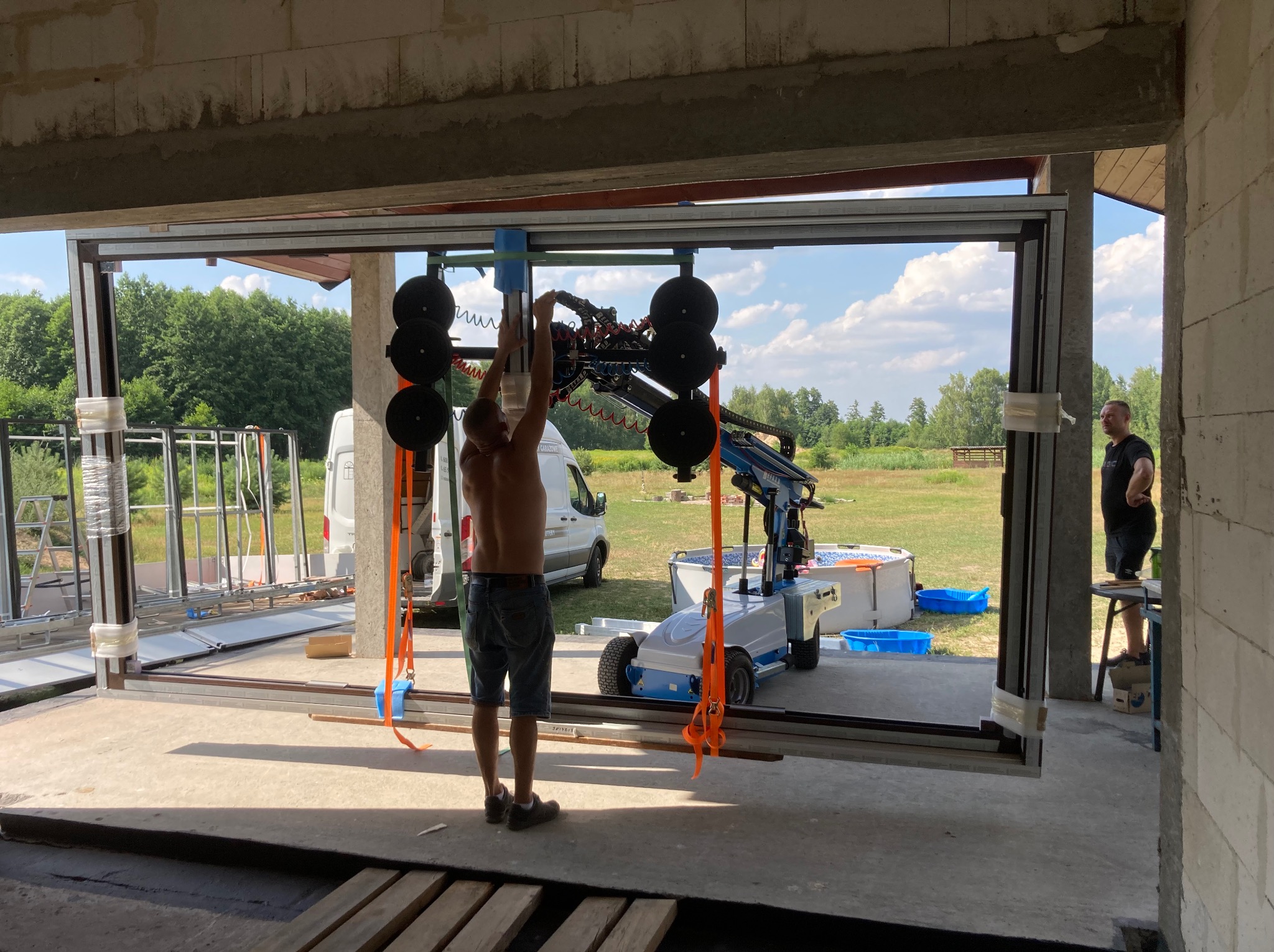 Montaż okien przesuwnych z użyciem specjalistycznego podnośnika próżniowego w surowym stanie budynku. Widoczny pracownik i częściowo zmontowana konstrukcja.