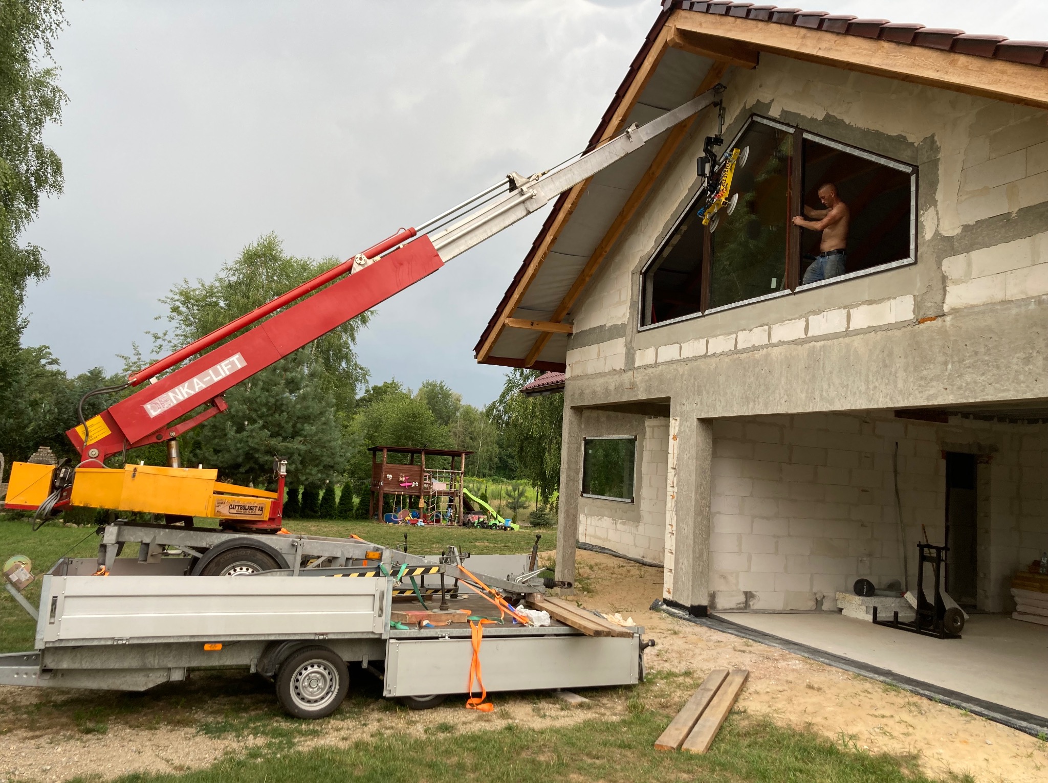 Montaż okna w surowym domu jednorodzinnym za pomocą podnośnika. Widoczny pracownik w oknie i podnośnik z przyssawkami. Dom w trakcie budowy.