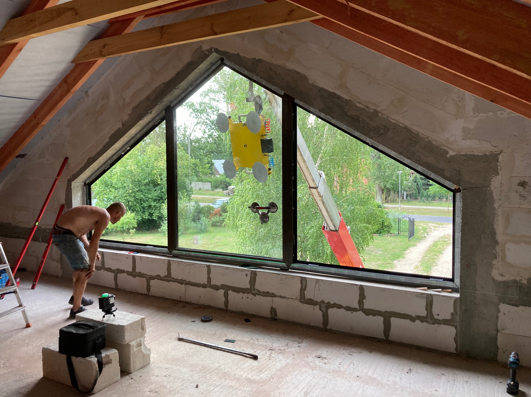Montaż okna trapezowego na poddaszu z użyciem przyssawki. Widoczny pracownik, laser krzyżowy i drabina. Surowy stan wykończenia wnętrza.