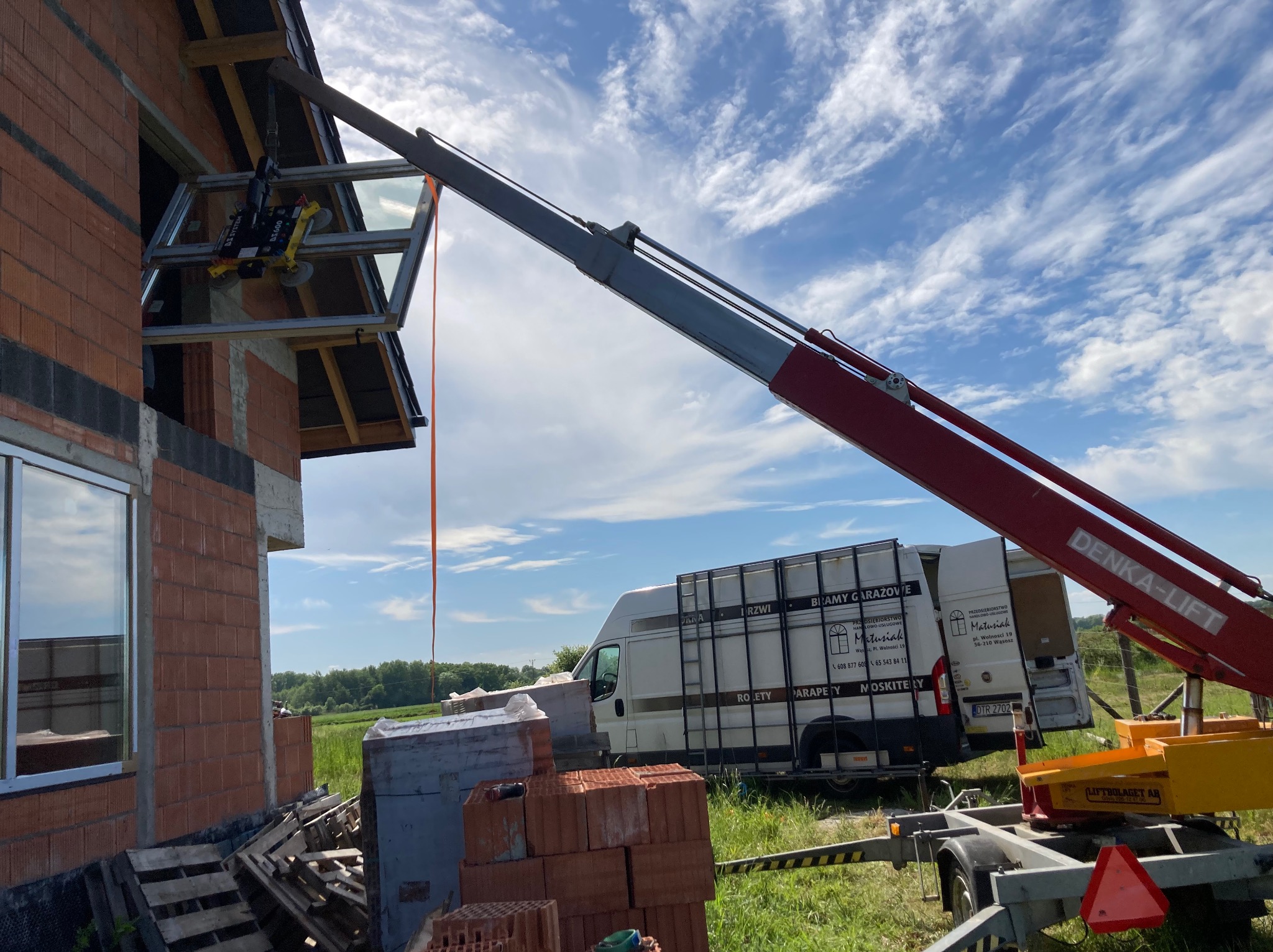 Montaż okna na budowie z użyciem dźwigu. Widoczny budynek w trakcie budowy, okno w ramie zawieszone na dźwigu, samochód dostawczy z reklamą firmy w tle.