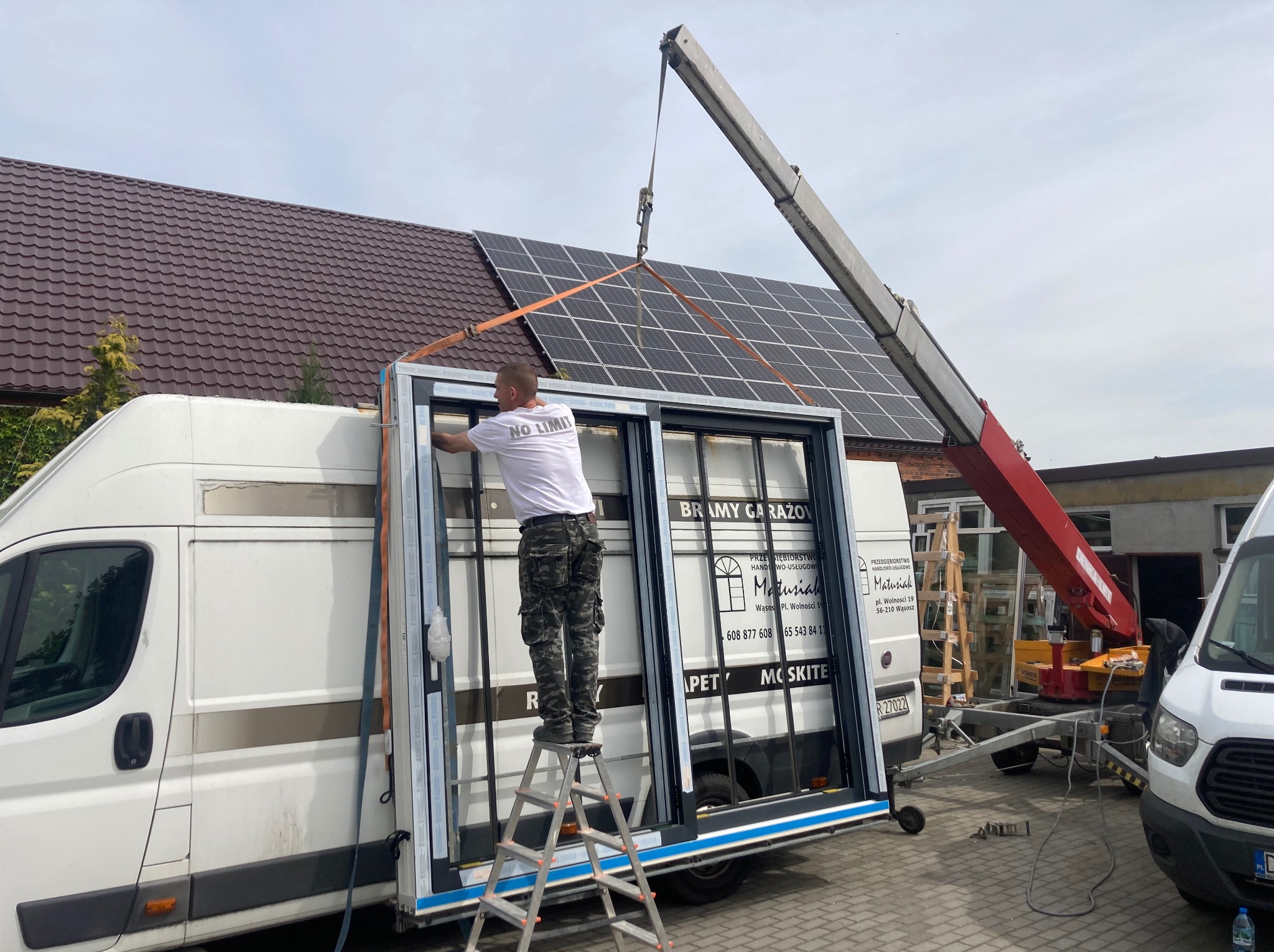 Montaż okna przesuwanego przez pracownika na drabinie, z użyciem dźwigu. Na dachu panele słoneczne. Widoczny samochód dostawczy z reklamą firmy.