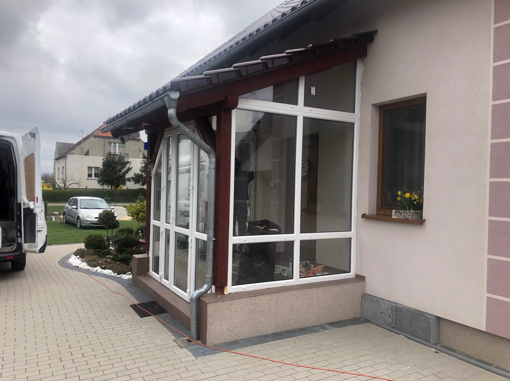 Zabudowa tarasu z białymi ramami okiennymi i brązowym dachem, widok z zewnątrz. Na ścianie budynku małe okno z kwiatami. Częściowo widoczny samochód dostawczy.