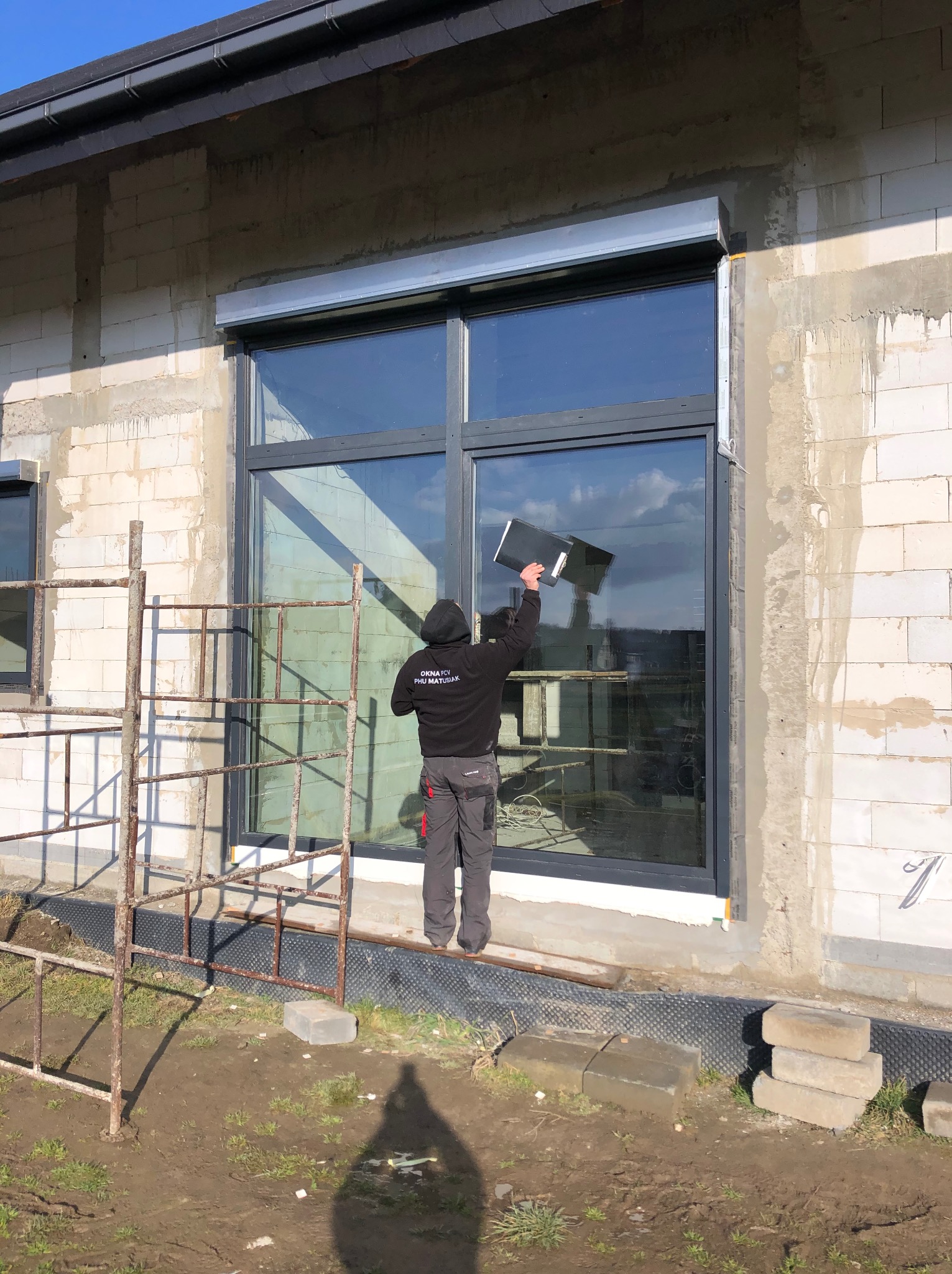 Montaż okna PCV z roletą zewnętrzną w surowym budynku. Pracownik wygładza folię ochronną na szybie. Widoczne rusztowanie i bloczki.