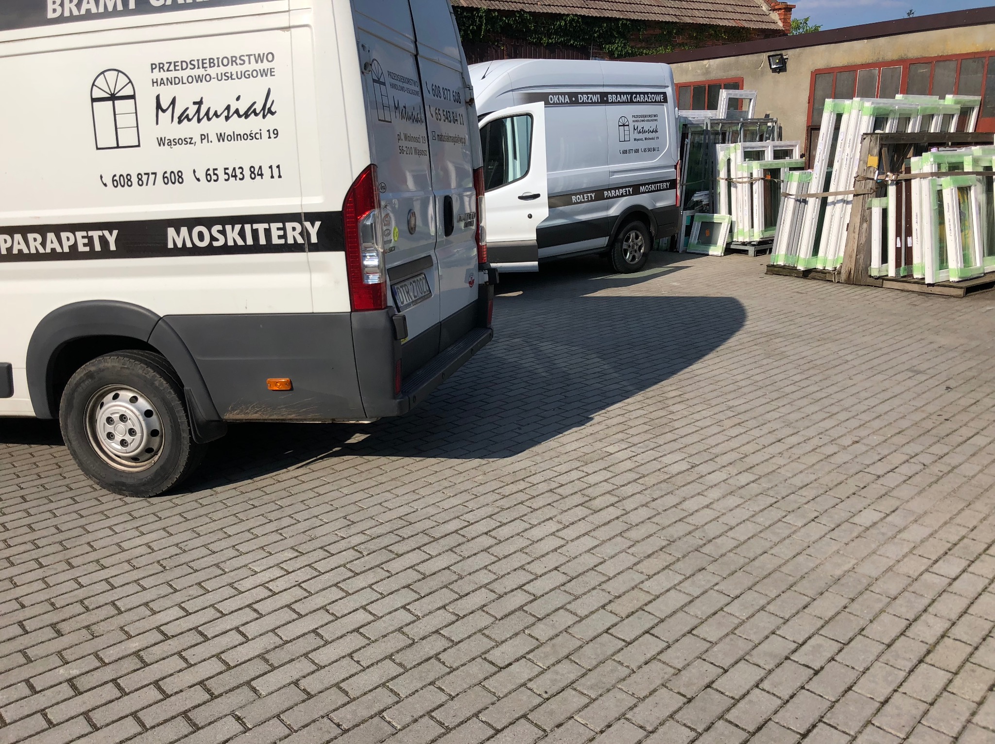 Dwa busy firmy Matusiak z reklamą okien, drzwi, rolet i moskitier na tle budynku i składowanych okien. Kostka brukowa na pierwszym planie.