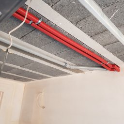 Fresh Art - Czerwone rury instalacji grzewczej przymocowane do sufitu z betonowych bloczków, szare rury obok. Ściana w tle przygotowana do malowania.