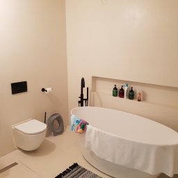Fresh Art - Minimalistyczna łazienka w beżach: owalna wanna z czarną armaturą, podwieszana toaleta i dywanik z frędzlami. W ścianie wnęka z kosmetykami.