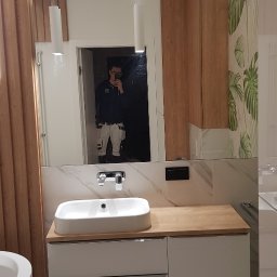 Fresh Art - Nowoczesna łazienka z umywalką nablatową, drewnianym blatem i szafką. W lustrze odbija się osoba robiąca zdjęcie. Widoczne oświetlenie i dekoracyjna tapeta.