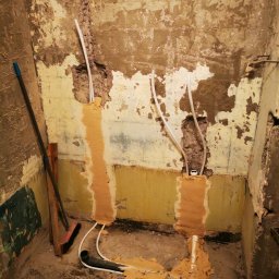 Fresh Art - Wnętrze łazienki w trakcie remontu: odsłonięte rury instalacji wodnej w ścianie, częściowo zamaskowane zaprawą. Widoczne ślady po starych płytkach i przygotowanie do dalszych prac.