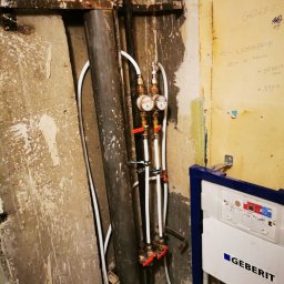 Fresh Art - Instalacja wodna z wodomierzami i zaworami, rury pionowe, białe rurki doprowadzające wodę, nieotynkowana ściana, stelaż podtynkowy Geberit w łazience w trakcie remontu.