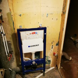 Fresh Art - Stelaż podtynkowy Geberit zamontowany w surowej łazience przed wykończeniem. Widoczne instalacje wodne i kanalizacyjne, ściany z resztkami starej farby i notatkami.