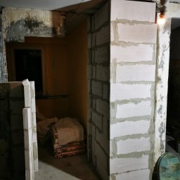 Fresh Art - Wznoszenie ścianki działowej z bloczków betonu komórkowego w surowym wnętrzu. Widoczne worki z zaprawą murarską. Żarówka oświetla miejsce pracy.