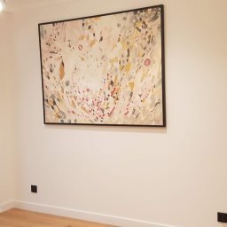 Fresh Art - Jasne wnętrze z abstrakcyjnym obrazem w czarnej ramie na białej ścianie. Widoczne drewniane panele dekoracyjne i nowoczesne lampy wiszące.