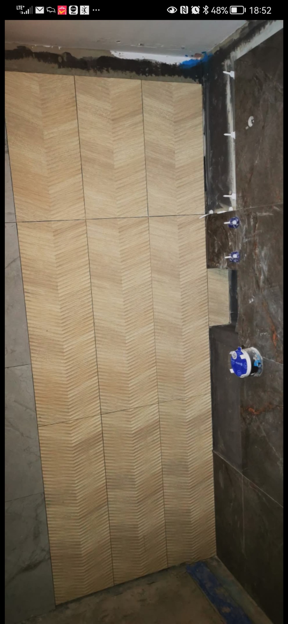 Ściana łazienki z ułożonymi płytkami w jodełkę, widoczne elementy instalacji wodnej. Częściowo wykończona ściana z ciemnymi płytkami.