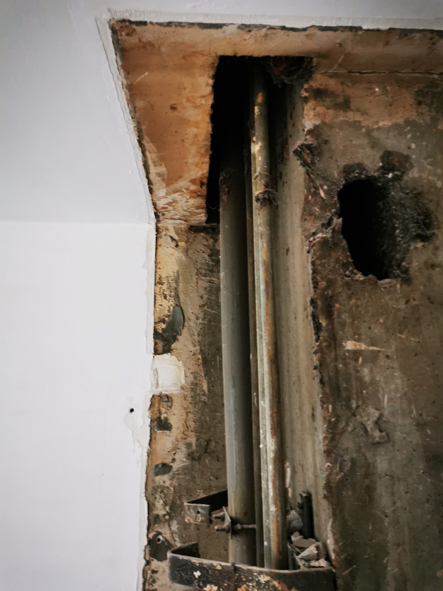 Odsłonięte rury w ścianie podczas remontu. Widoczne fragmenty starego tynku i świeżo pomalowana ściana. Metalowe uchwyty podtrzymujące rury.