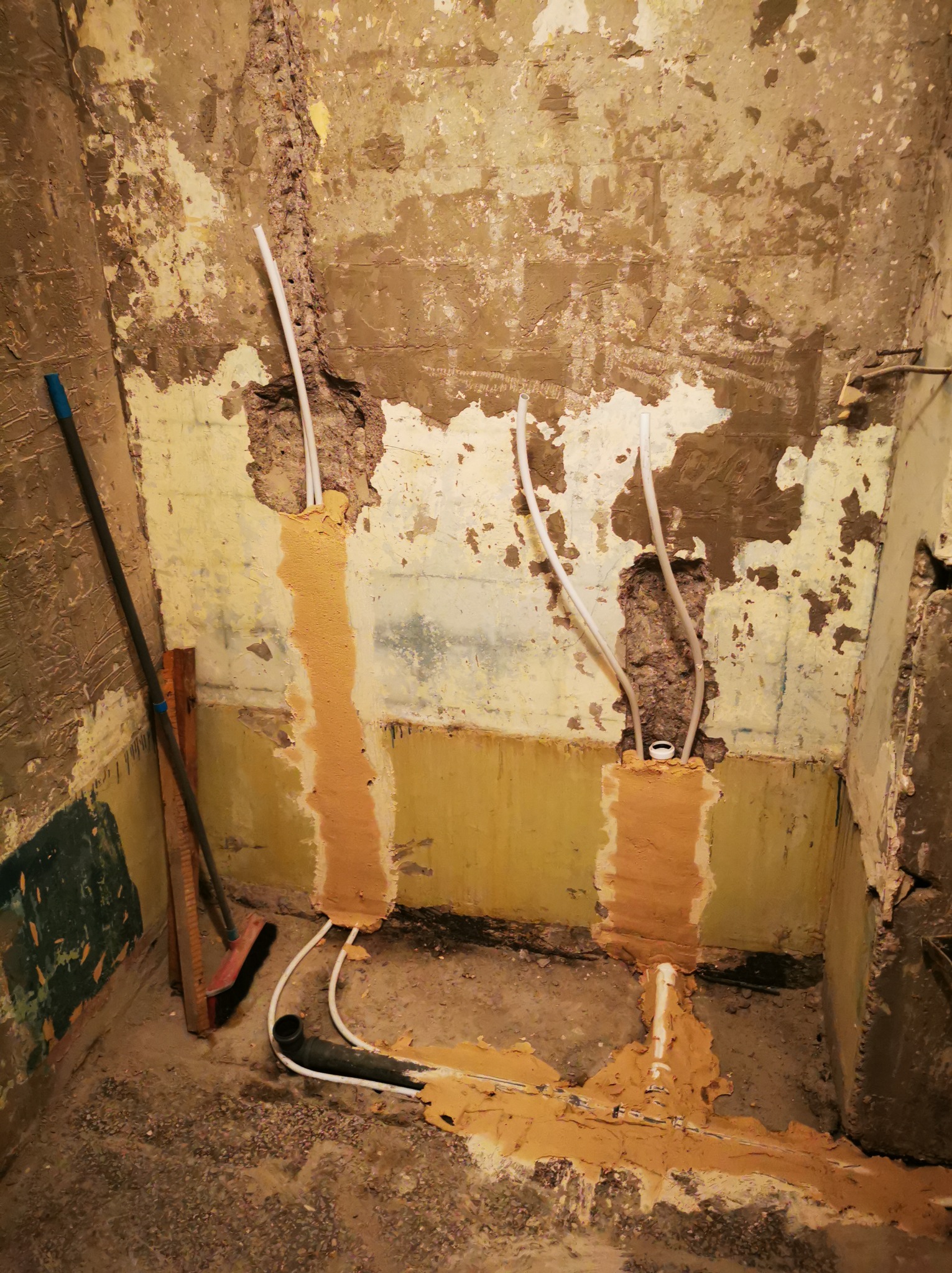 Wnętrze łazienki w trakcie remontu: odsłonięte rury instalacji wodnej w ścianie, częściowo zamaskowane zaprawą. Widoczne ślady po starych płytkach i przygotowanie do dalszych prac.