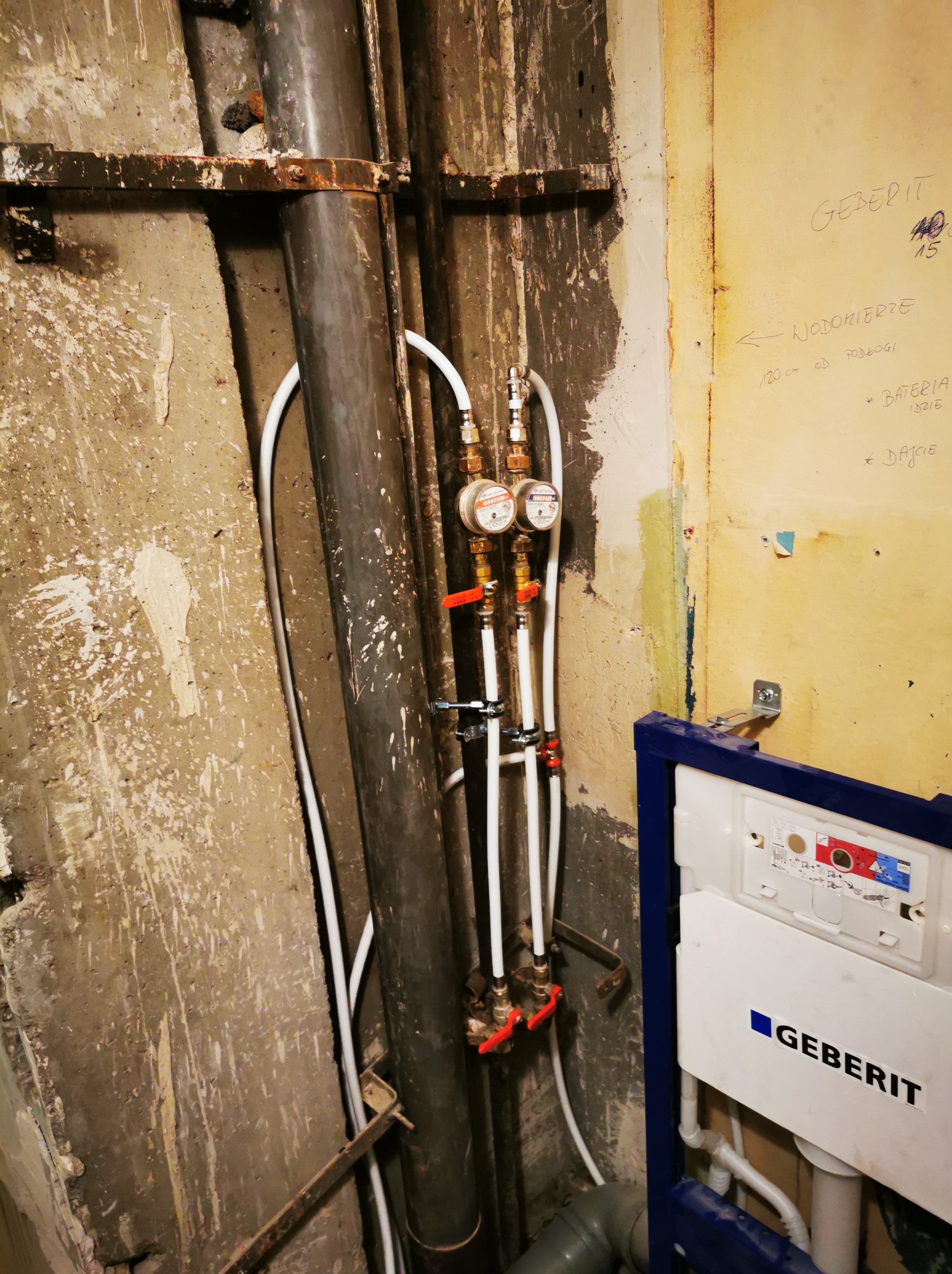 Instalacja wodna z wodomierzami i zaworami, rury pionowe, białe rurki doprowadzające wodę, nieotynkowana ściana, stelaż podtynkowy Geberit w łazience w trakcie remontu.