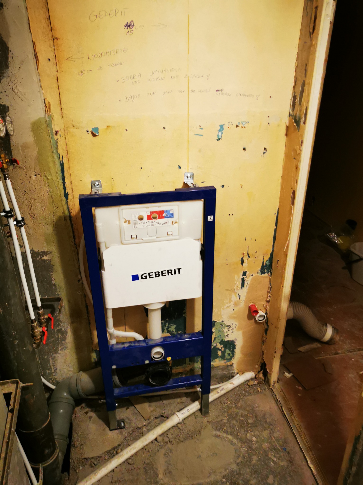 Stelaż podtynkowy Geberit zamontowany w surowej łazience przed wykończeniem. Widoczne instalacje wodne i kanalizacyjne, ściany z resztkami starej farby i notatkami.
