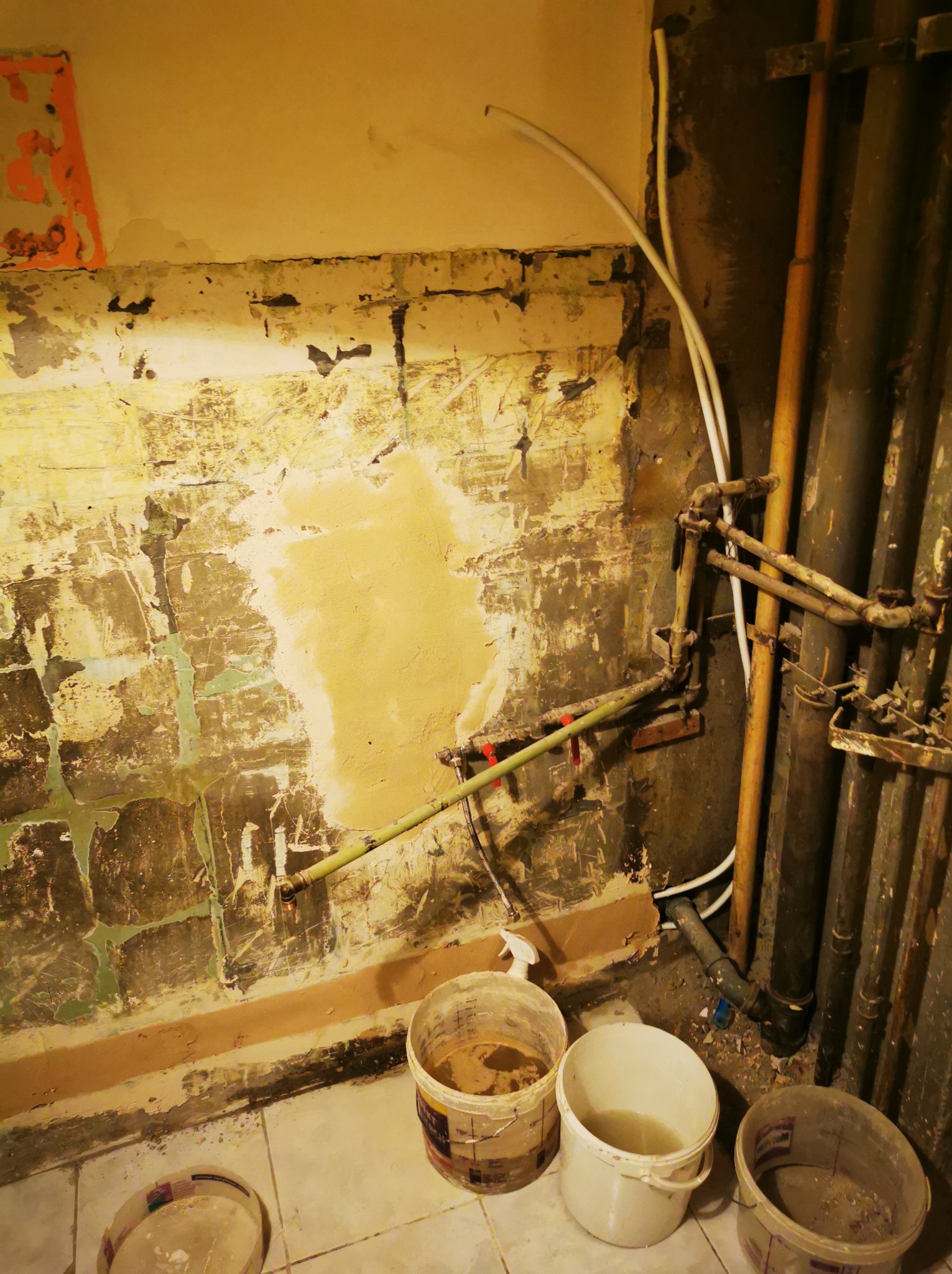 Ściana w trakcie remontu z widocznymi rurami, fragmentami starej farby i nałożoną gładzią. Na podłodze stoją wiadra z resztkami farby.
