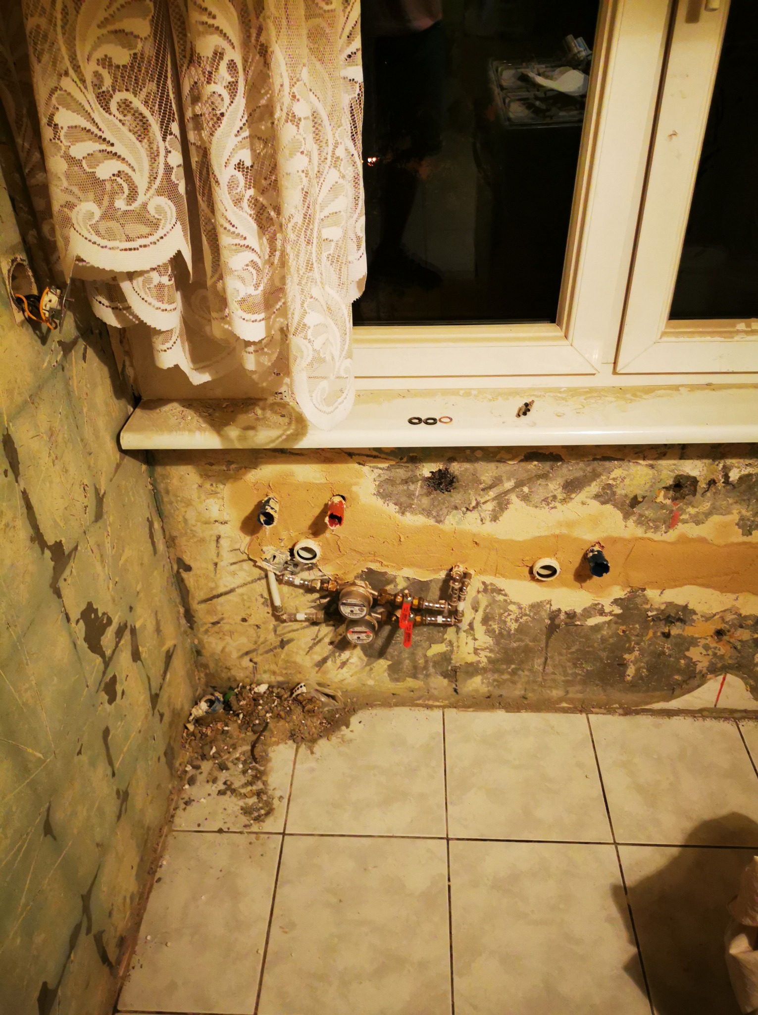 Fragment łazienki w trakcie remontu: odsłonięte rury, liczniki, stara glazura, okno z koronkową firanką. Widoczne ślady po kuciu w ścianie.