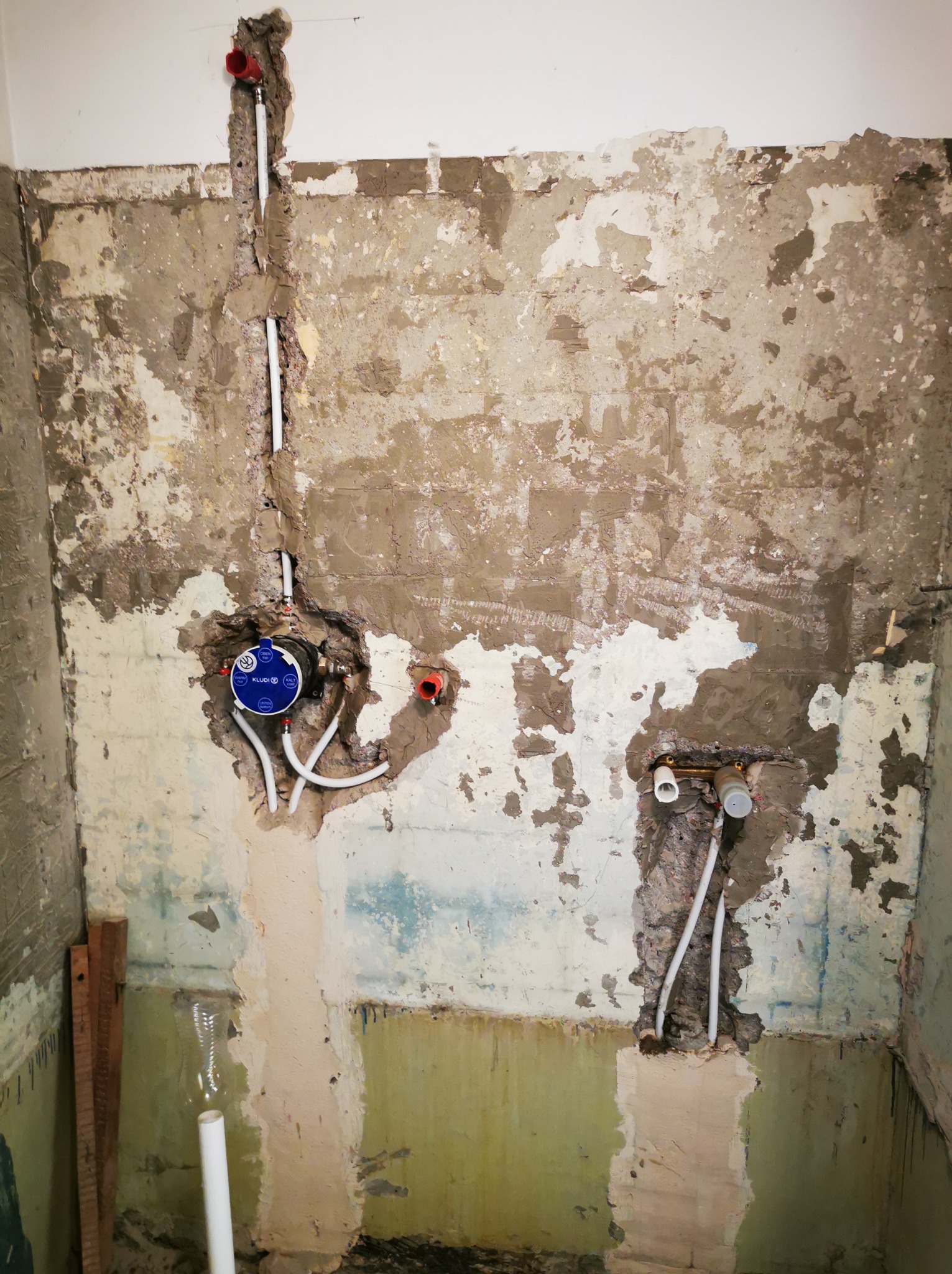 Ściana łazienki w trakcie remontu z widocznymi instalacjami wodnymi i fragmentami starej farby. Widoczne rury i elementy montażowe.