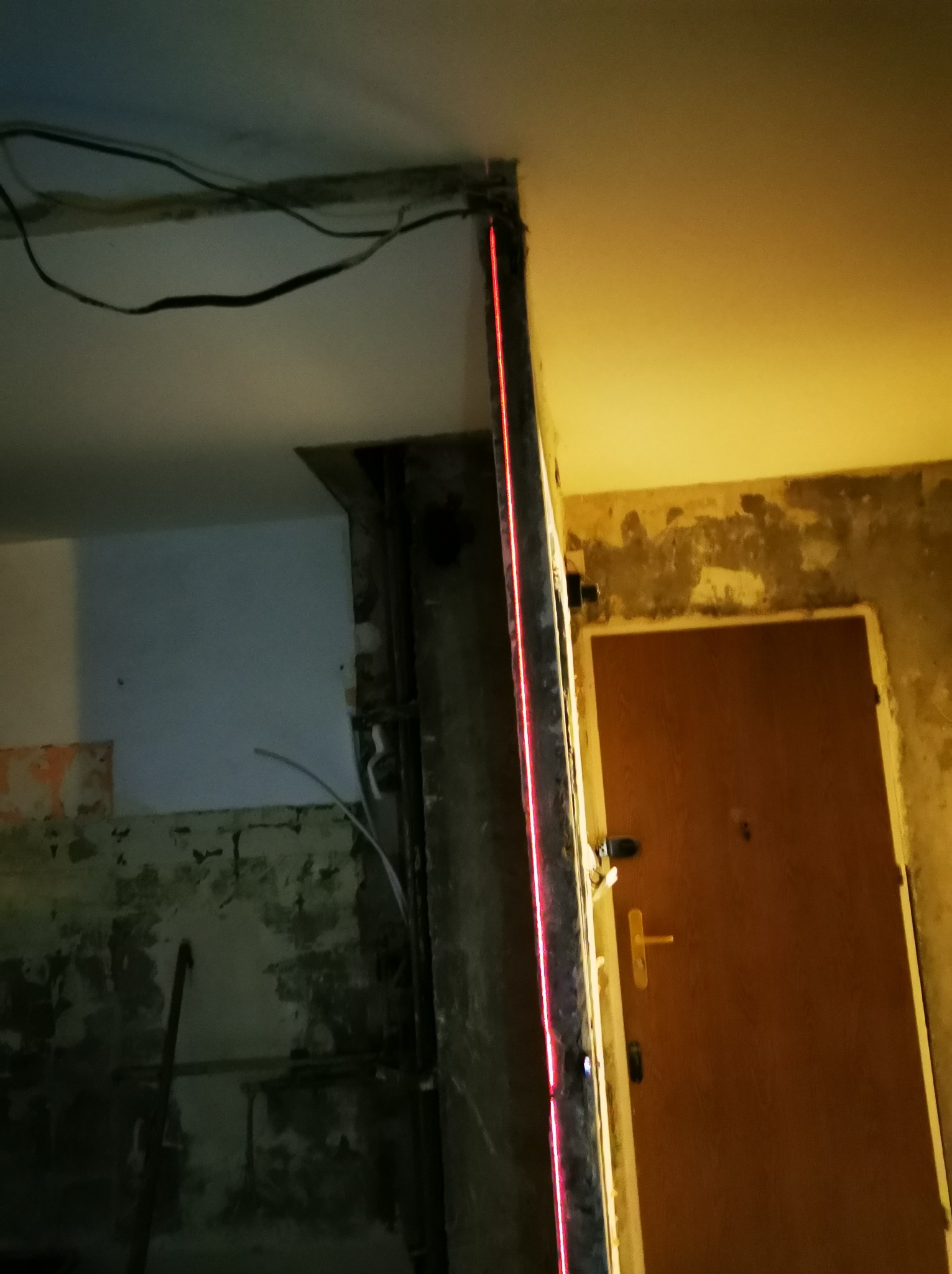 Remont pokoju: laserowy pomiar pionu na ścianie z widocznymi śladami starej farby, przygotowanie do montażu ościeżnicy. Widoczne przewody elektryczne.