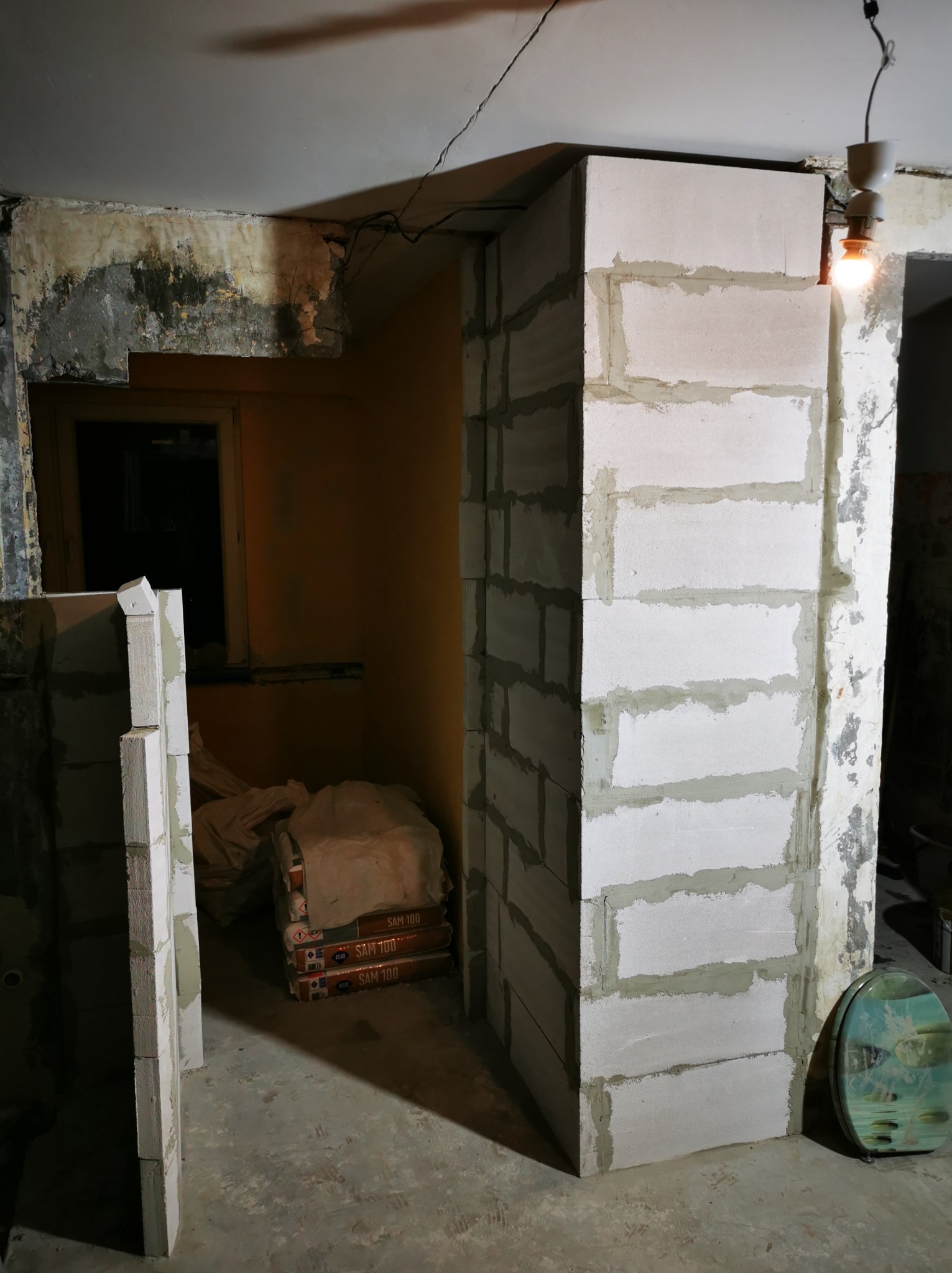Wznoszenie ścianki działowej z bloczków betonu komórkowego w surowym wnętrzu. Widoczne worki z zaprawą murarską. Żarówka oświetla miejsce pracy.