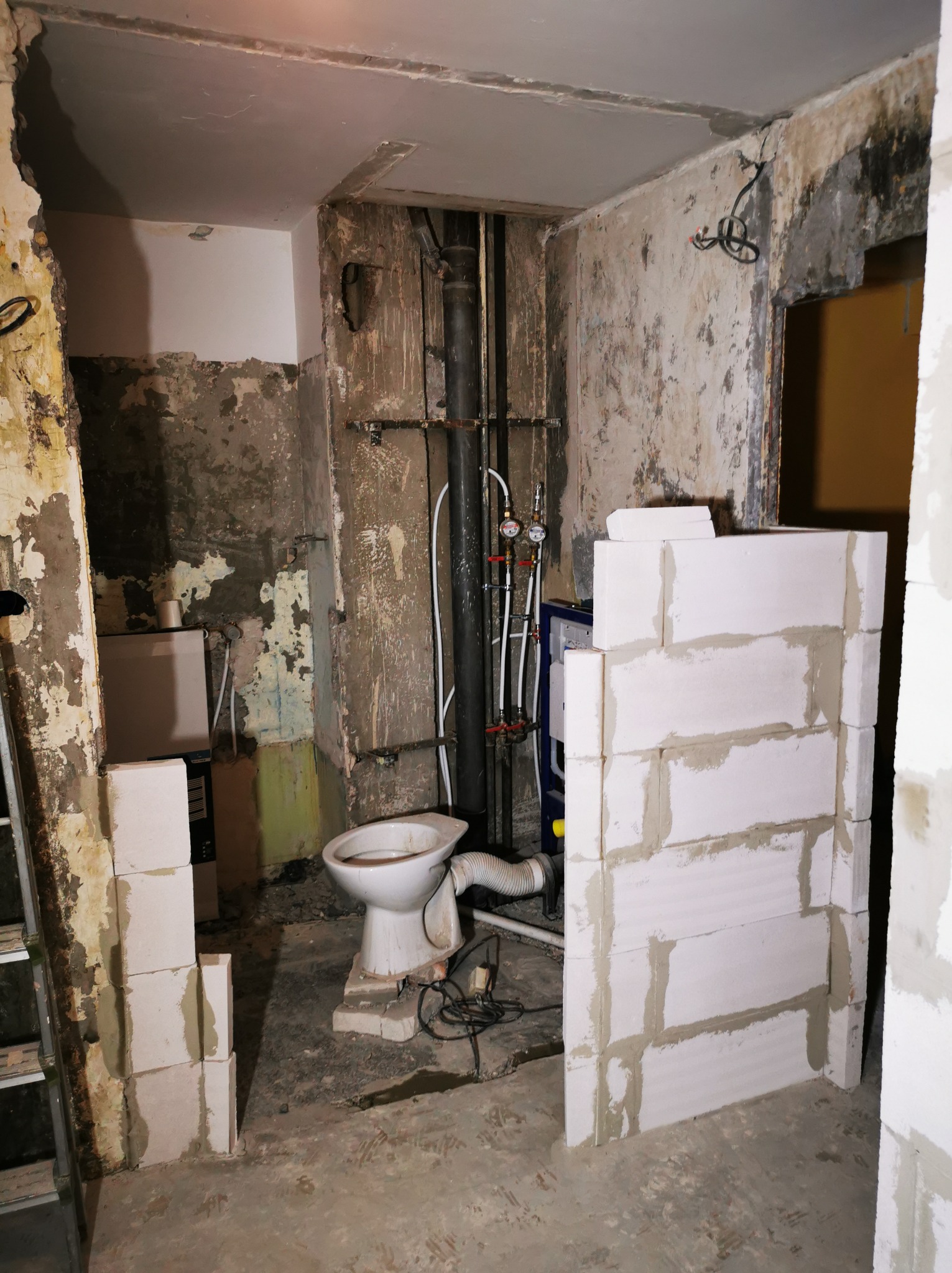 Remont łazienki w trakcie: murowanie ścianki działowej z bloczków, widoczne instalacje, stara toaleta. Surowe ściany z resztkami farby i tynku. Prace murarskie.