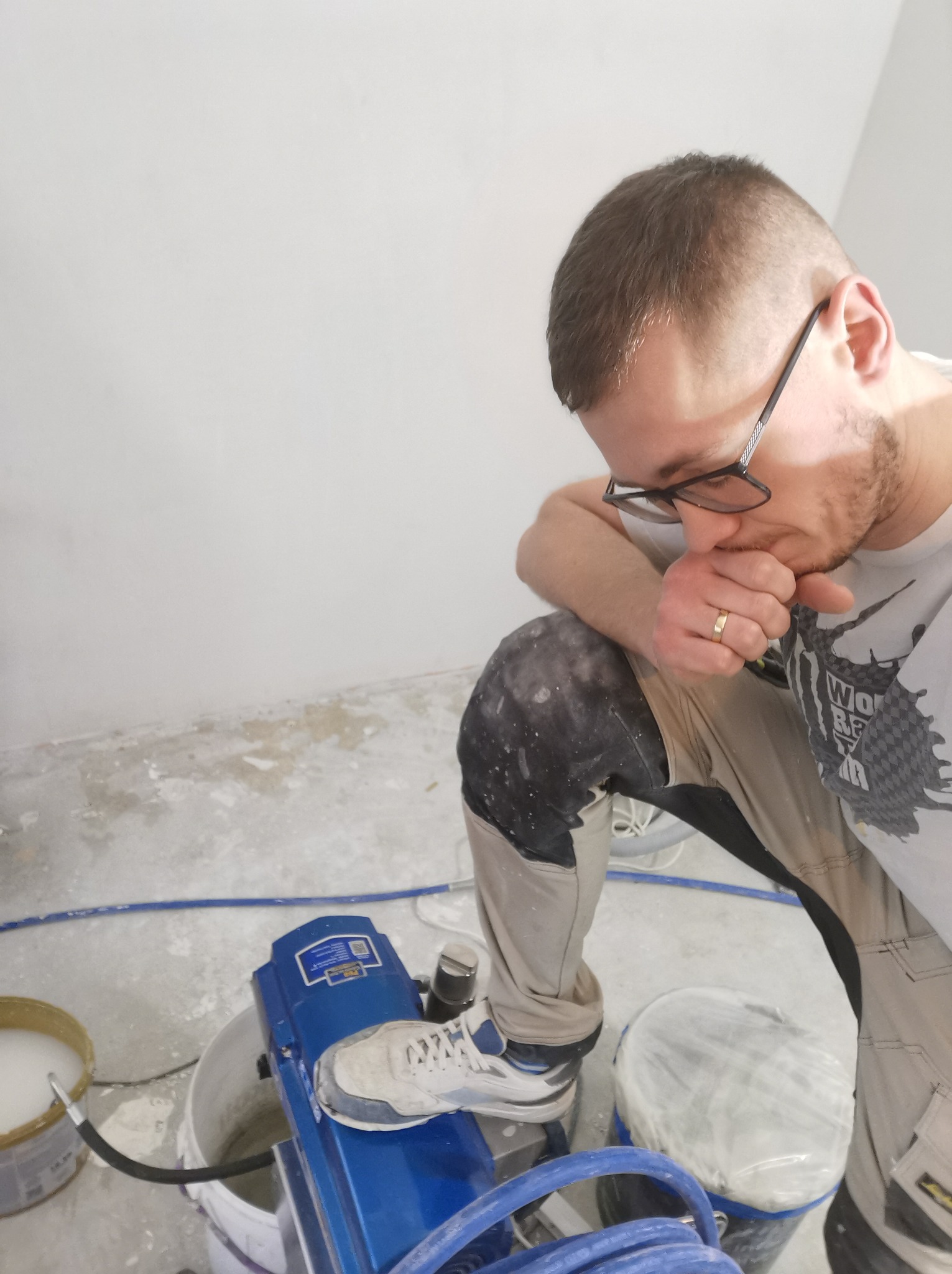 Pracownik w okularach, w trakcie pracy z agregatem malarskim, oparty na kolanie, z widocznym wiaderkiem z farbą i niepomalowaną ścianą w tle.