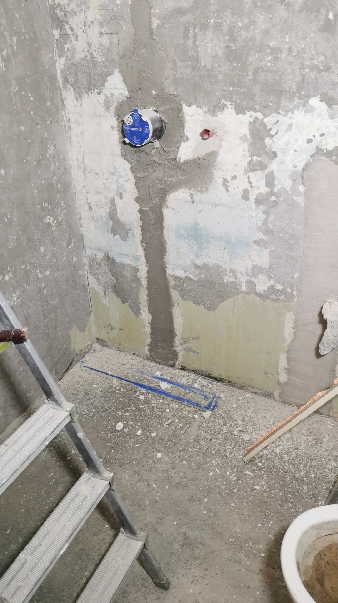 Narożnik łazienki w trakcie remontu: surowe ściany z fragmentami starej farby, zamontowany zawór Kludi, drabina i narzędzia na podłodze. Widoczne ślady po szpachlowaniu.