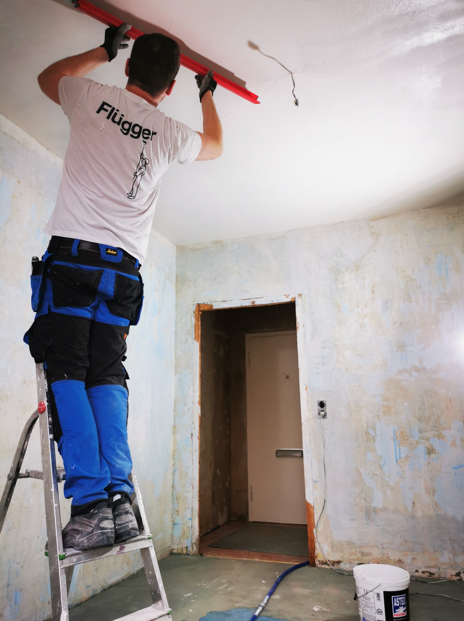 Pracownik na drabinie, z wałkiem malarskim, maluje sufit w pomieszczeniu w trakcie remontu. Widoczne ślady po starych powłokach na ścianach.