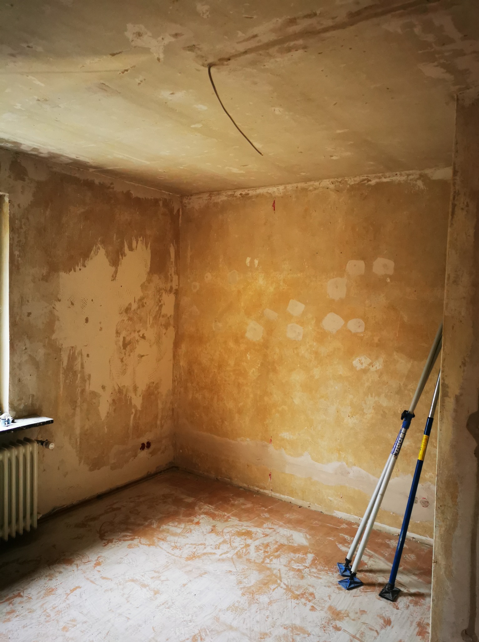 Pokój w trakcie remontu: ściany z widocznymi śladami po szpachlowaniu, podłoga zabezpieczona folią, podpory budowlane w rogu. Widoczne okno i grzejnik.