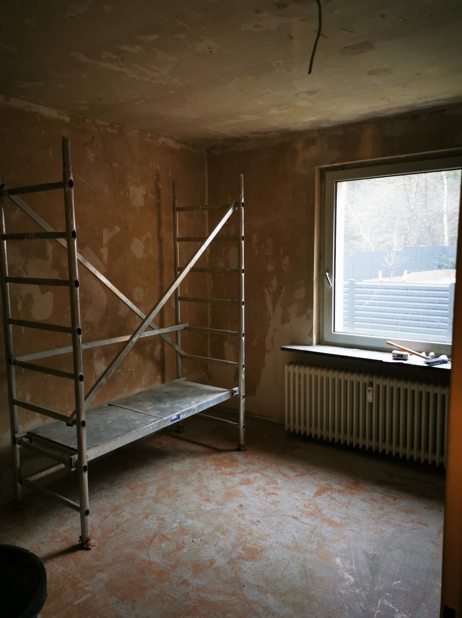 Pokój w trakcie przygotowań do remontu. Widoczne rusztowanie, ściany z resztkami starej farby, okno z grzejnikiem i narzędzia na parapecie. Podłoga częściowo oczyszczona.