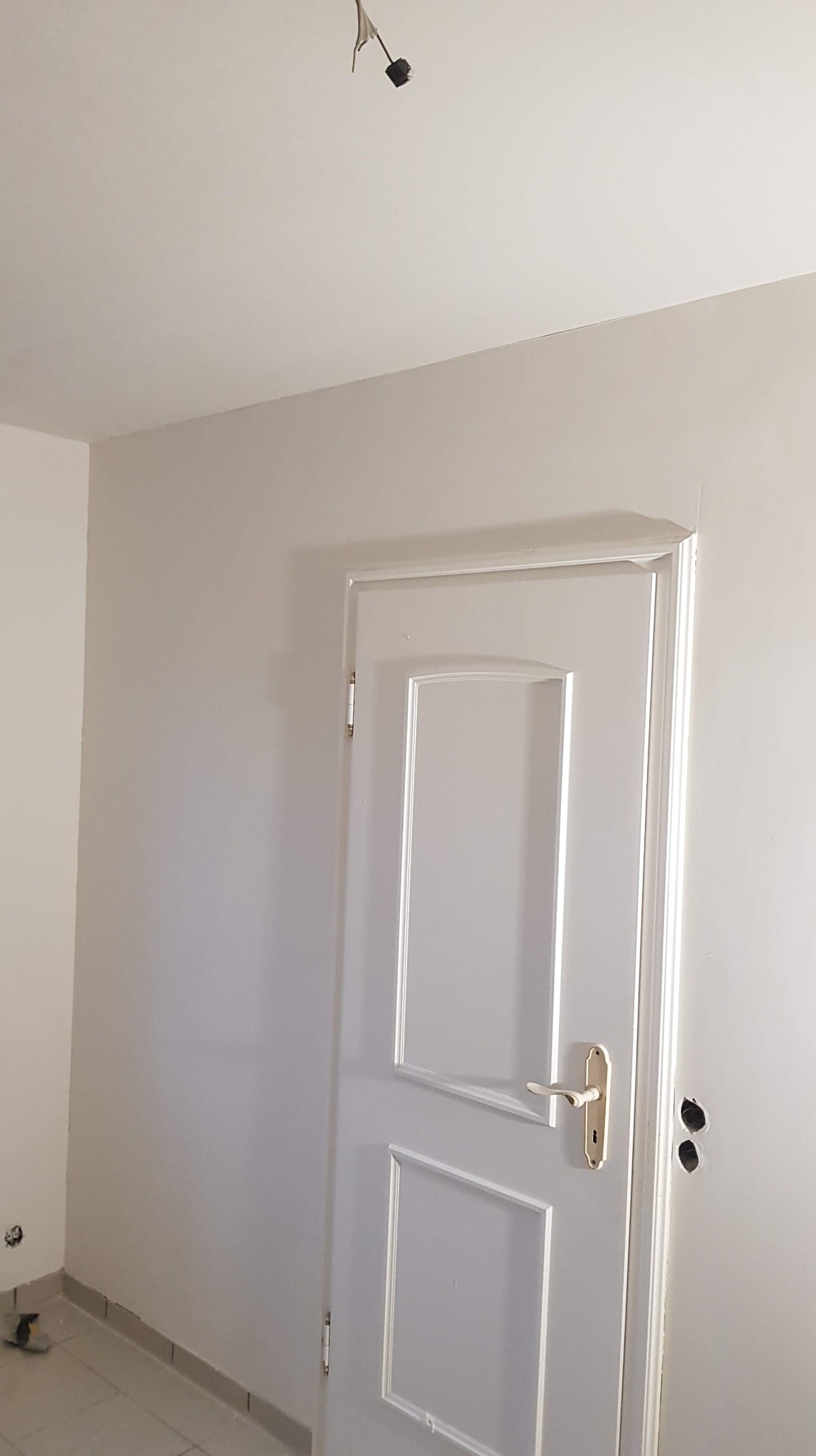 Białe, odświeżone wnętrze po malowaniu: ściana, drzwi z ozdobnymi ramkami i złotą klamką. Widoczne otwory po gniazdkach elektrycznych i wystający kabel od lampy.