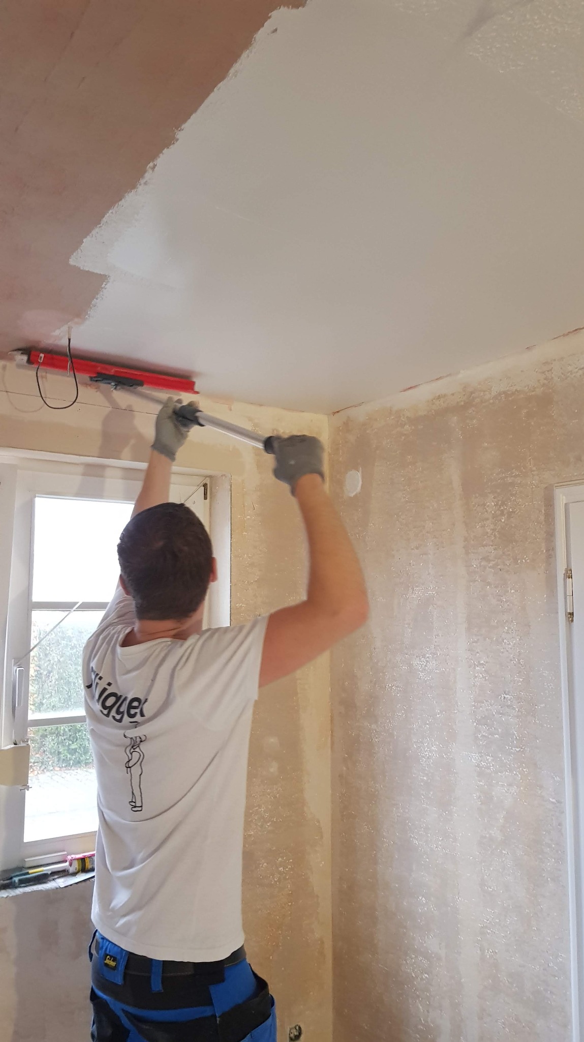 Pracownik w trakcie szpachlowania sufitu za pomocą szpachli z długim uchwytem. Widoczne częściowo wyszpachlowane fragmenty sufitu i przygotowane ściany.