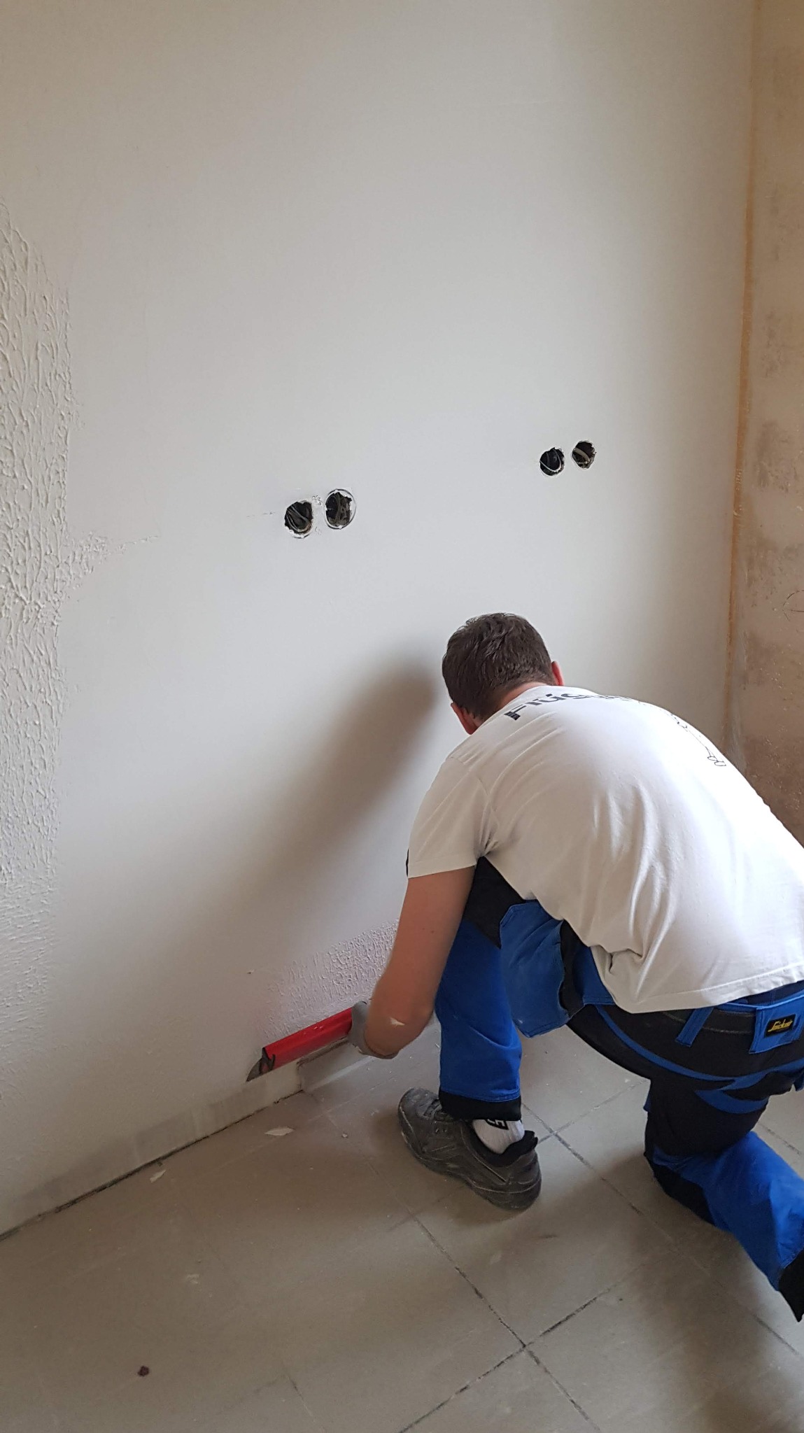 Pracownik w trakcie szpachlowania ściany w pomieszczeniu z widocznymi puszkami elektrycznymi i fragmentem ściany z fakturą. Używa czerwonej szpachelki.